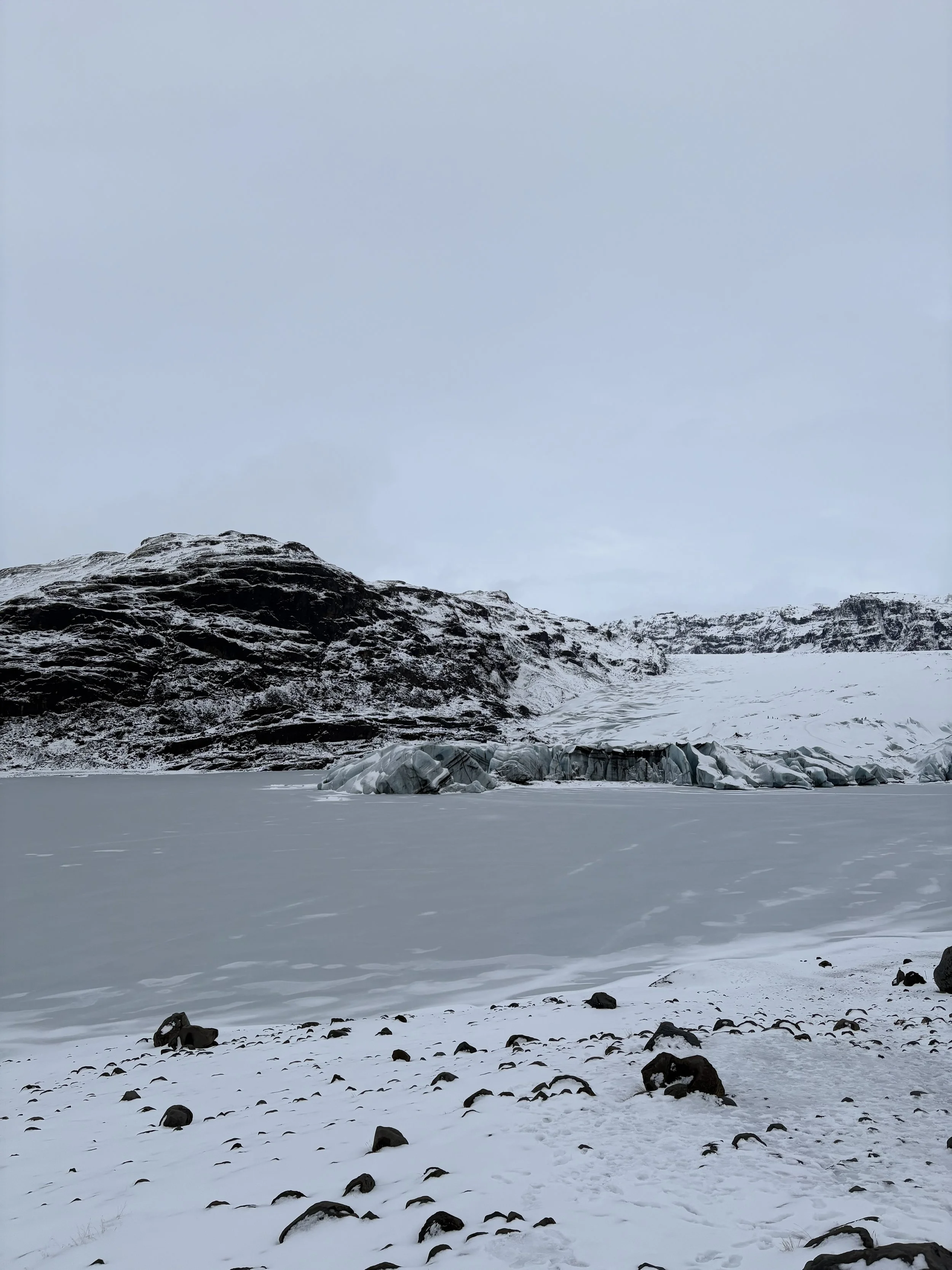 Snow-covered landscape with rocky hills and glaciers under a cloudy sky.
