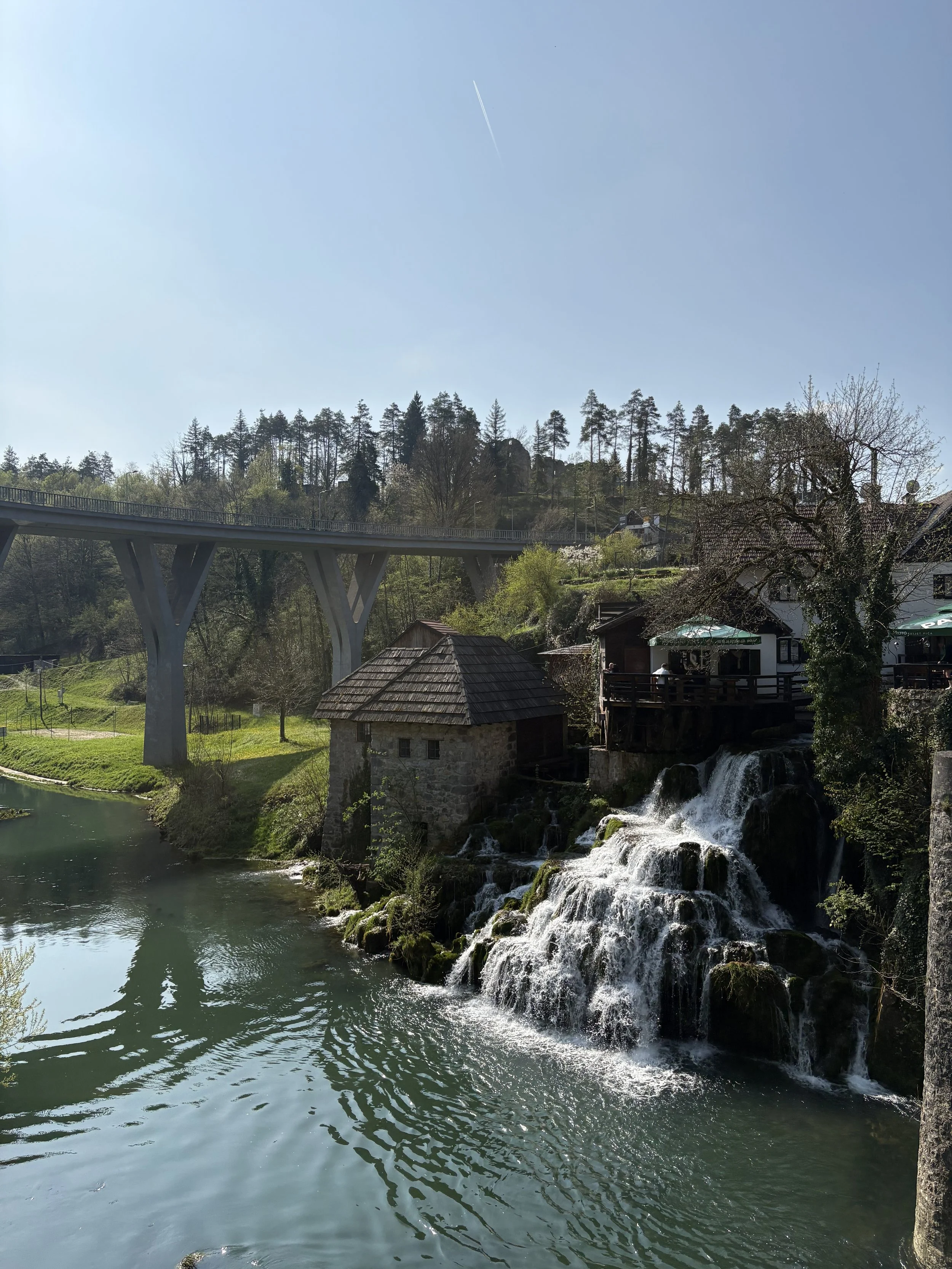 A scenic landscape featuring a waterfall cascading into a river, a bridge towering over the water, and a hillside with trees and houses under a clear blue sky.