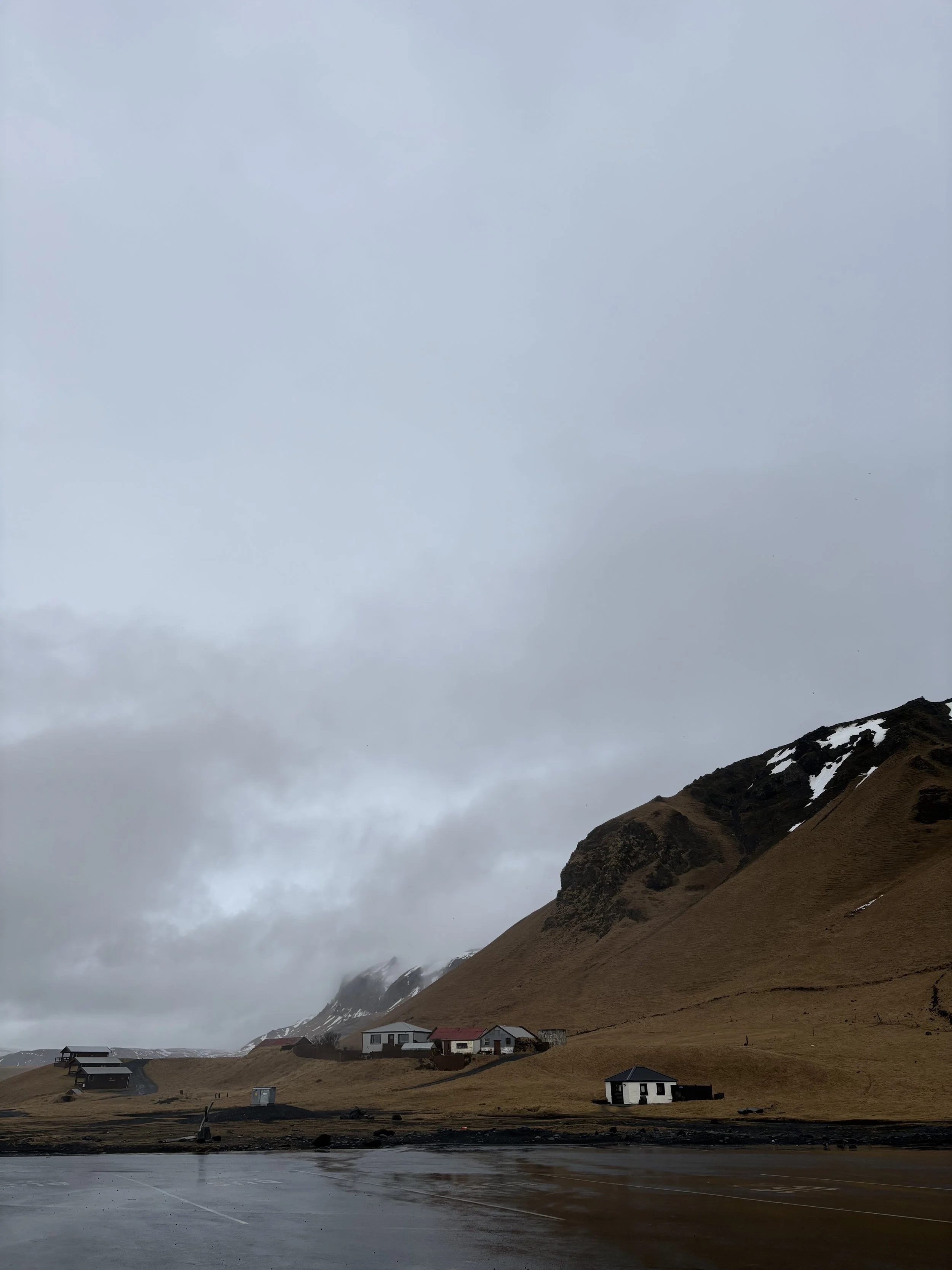 A small village at the base of a mountain with patches of snow, houses with white walls and dark roofs, a body of water in the foreground, and cloudy sky overhead.