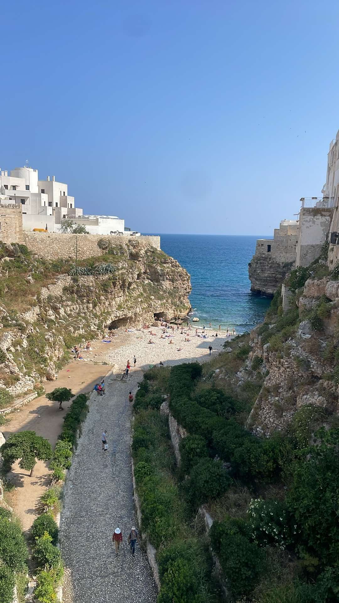 A scenic view of a small beach cove surrounded by rocky cliffs and white Mediterranean-style buildings on a sunny day.