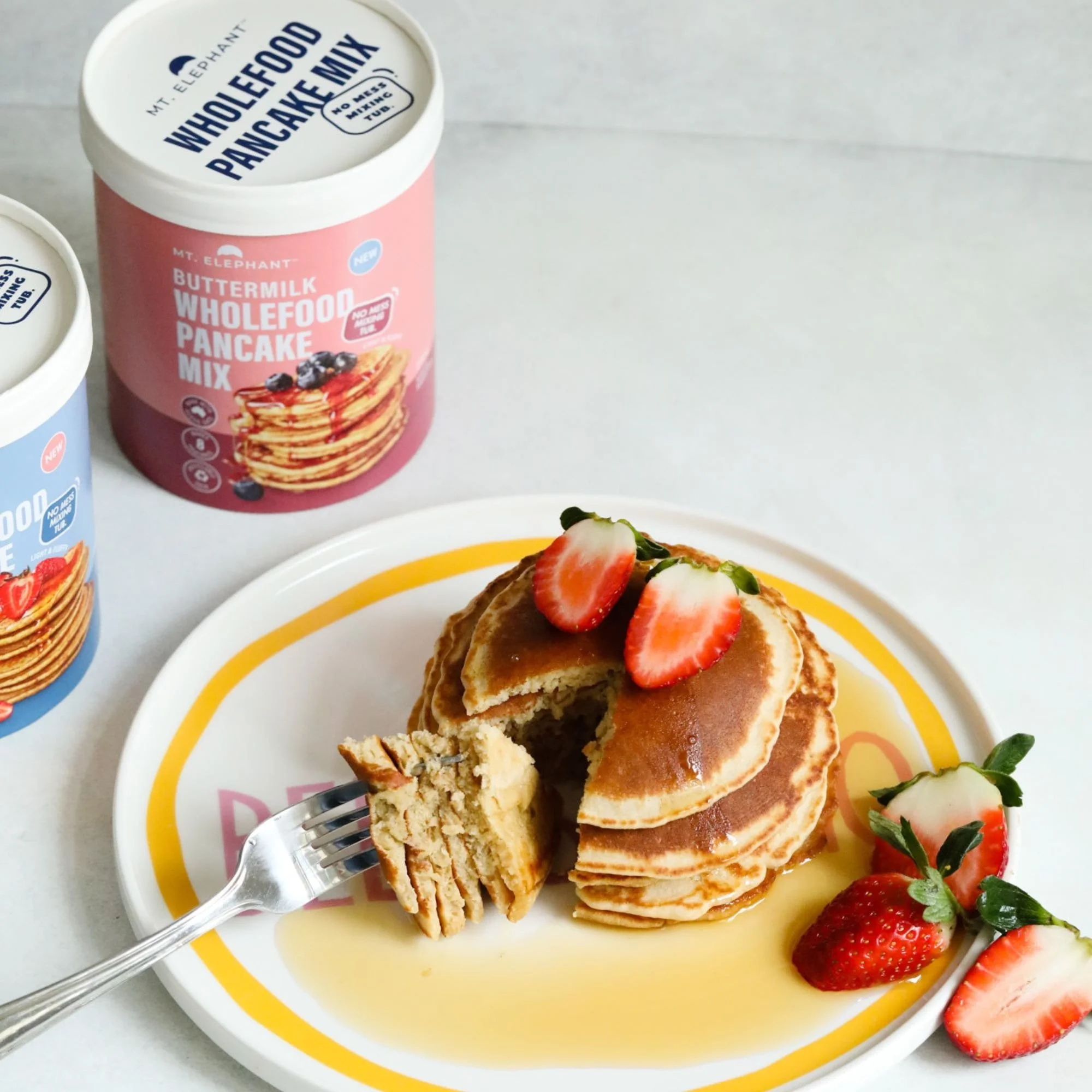 Plate of pancakes topped with sliced strawberries and syrup, with a container of Wholefood Pancake Mix and strawberries in the background.
