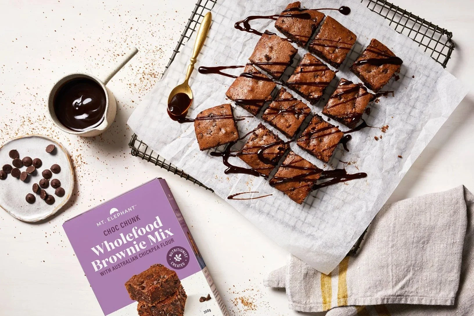 Chocolate brownies drizzled with chocolate sauce on a wire cooling rack, with a cup of melted chocolate, chocolate chips, and a purple box of wholefood brownie mix nearby.