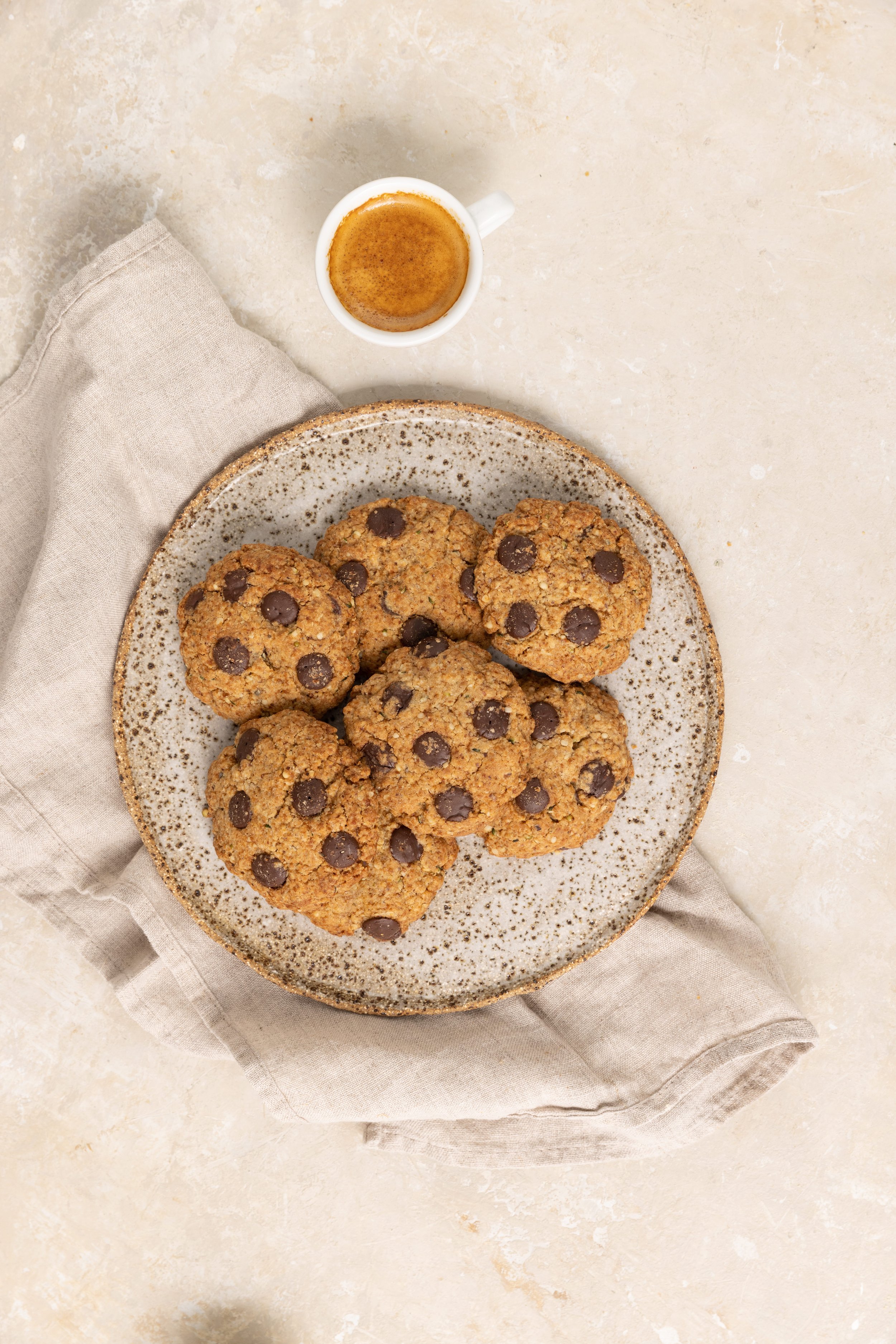 A plate of oatmeal cookies with chocolate chips on a speckled ceramic plate, next to a cup of espresso and a beige cloth napkin on a light-colored surface.