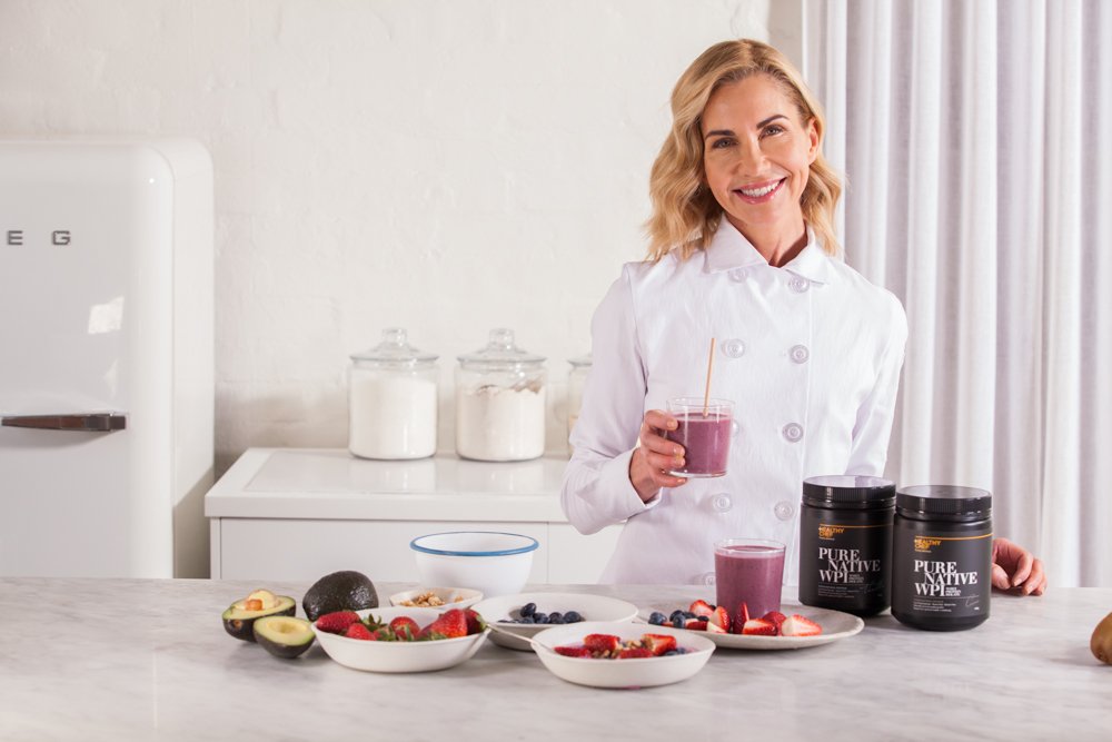 A woman in a white chef's coat smiling and holding a glass of purple smoothie in a bright kitchen with white countertops, bowls of fruit, and dietary supplements.