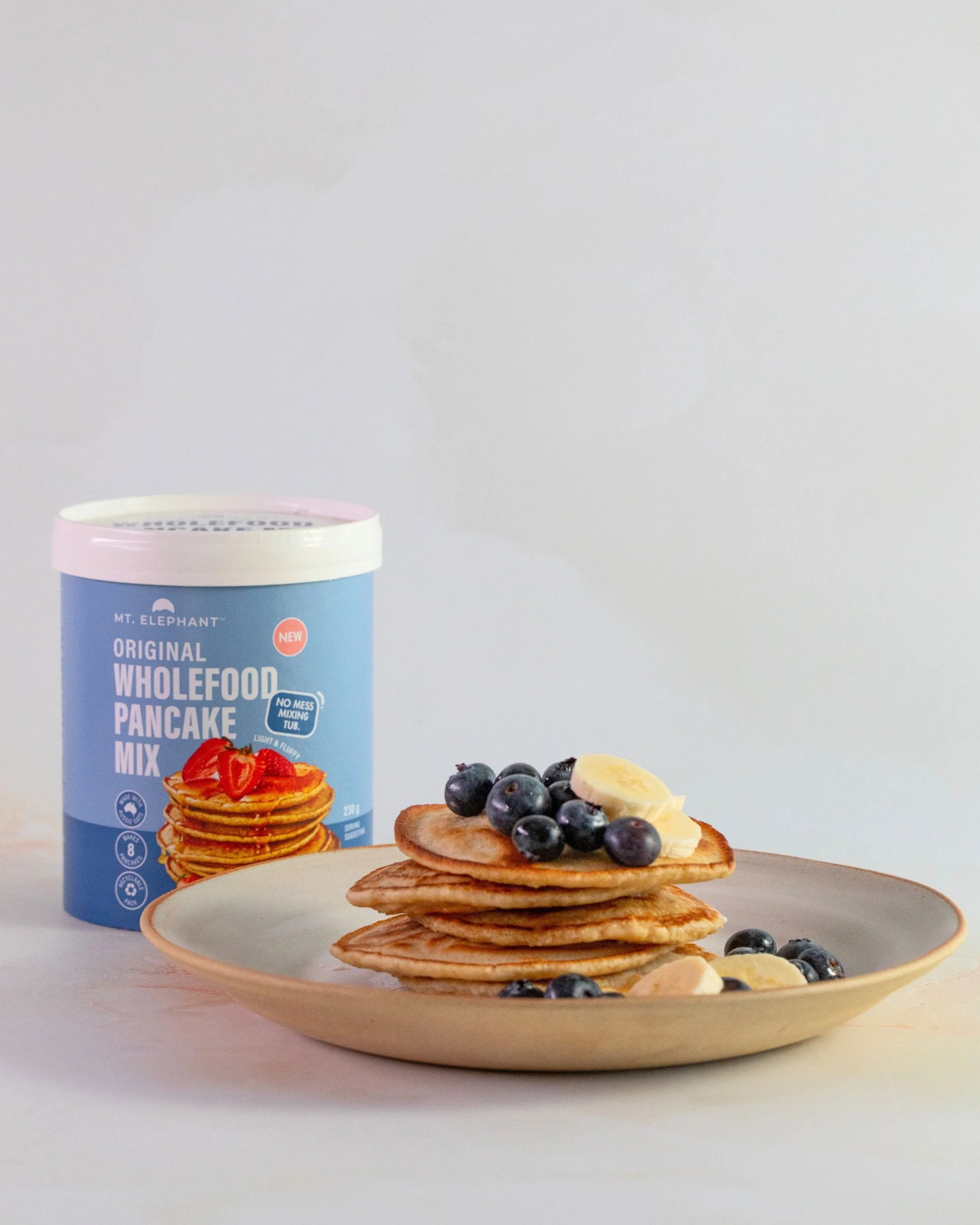 A plate of pancakes topped with blueberries and banana slices, with more blueberries scattered around, and a can of Mt. Elephant pancake mix in the background.