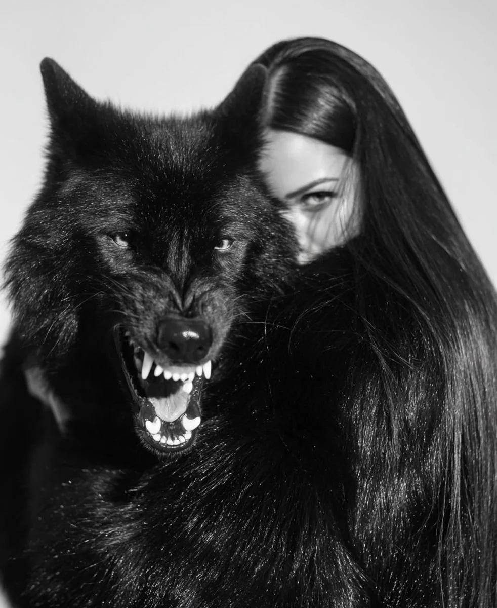 A woman with long, straight black hair partially hidden behind a snarling black wolf with sharp teeth, in a black-and-white photograph.