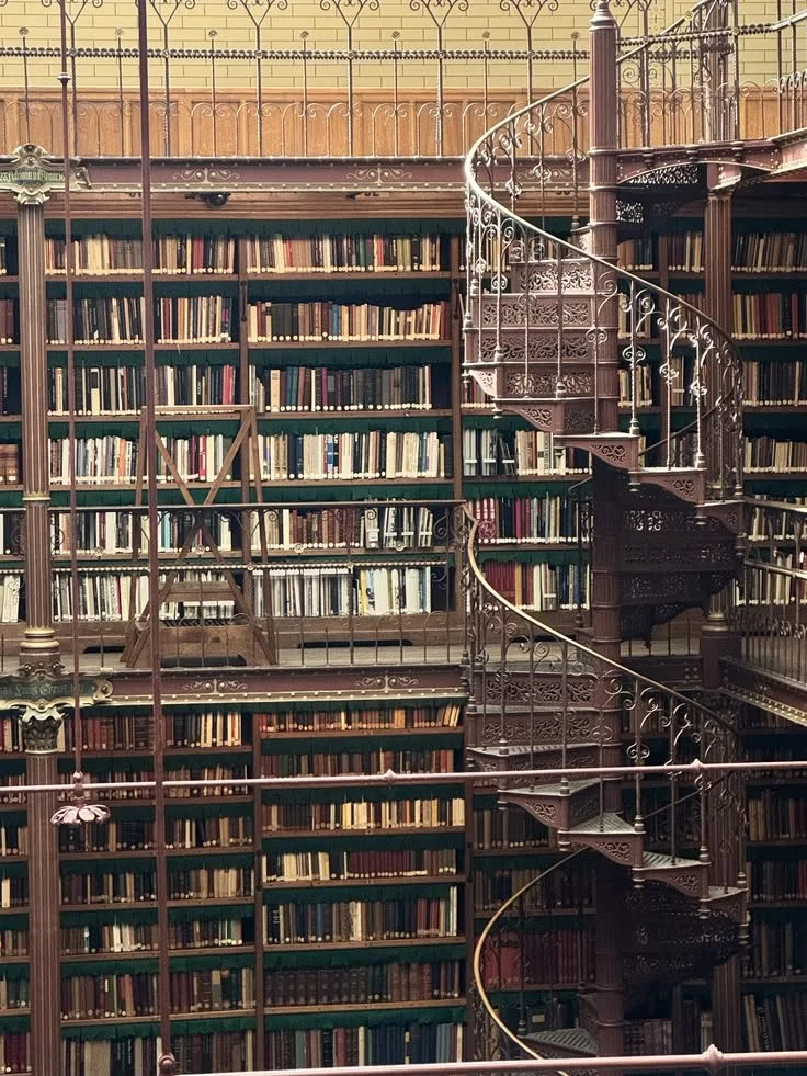 Multiple bookshelves filled with numerous books, with a decorative metal spiral staircase in the center of the photo, inside a vintage library or bookstore.