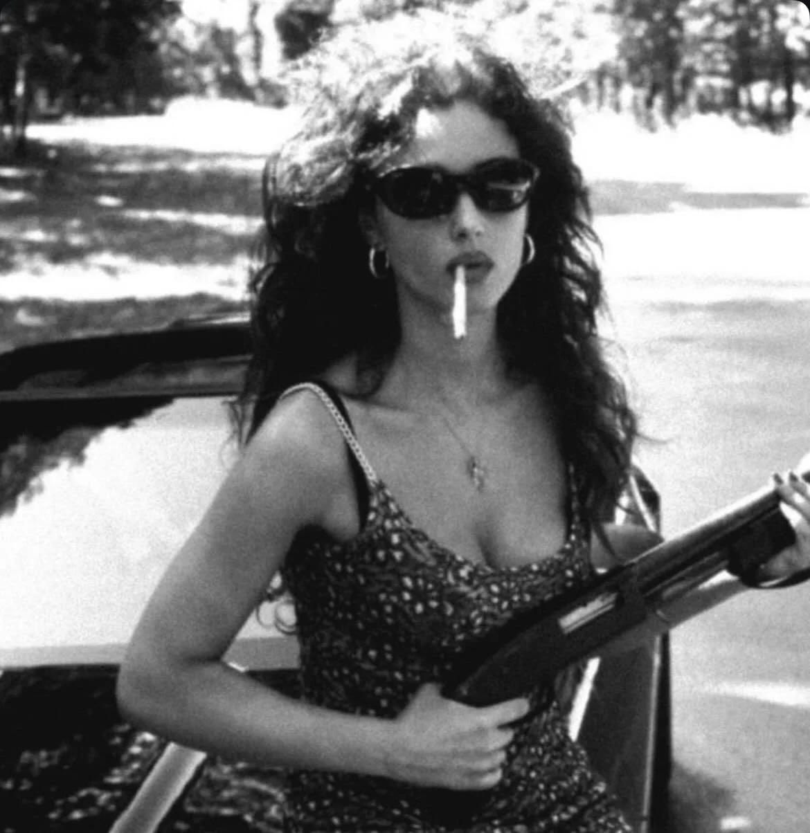 A woman with curly hair wearing sunglasses and hoop earrings, smoking a cigarette and holding a shotgun outdoors next to a car in a wooded area.