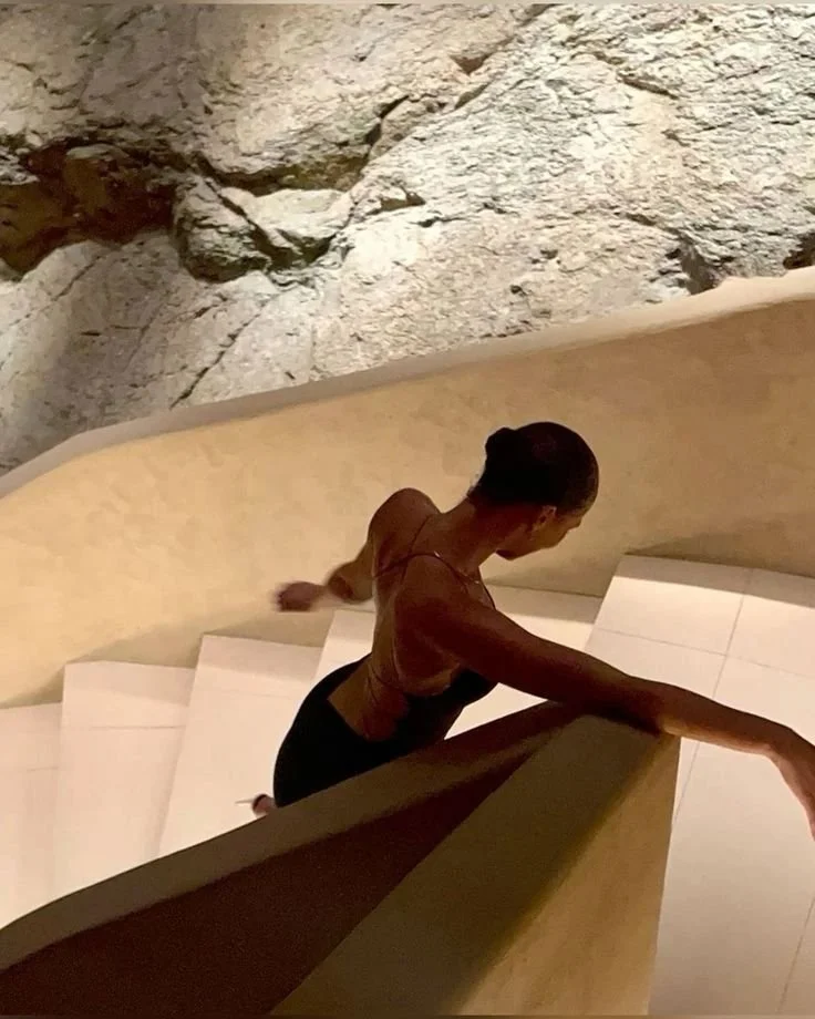 A woman with dark hair in a bun is ascending curving stairs with a carved stone wall in the background.