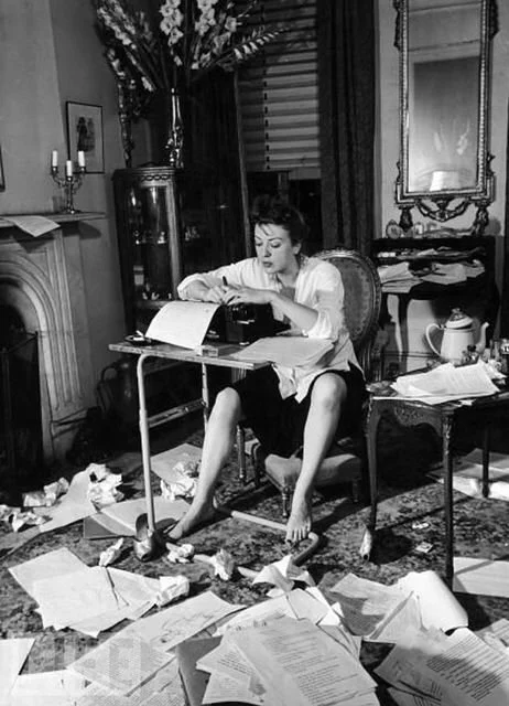 Woman working at a cluttered desk with papers on the floor in a vintage living room.