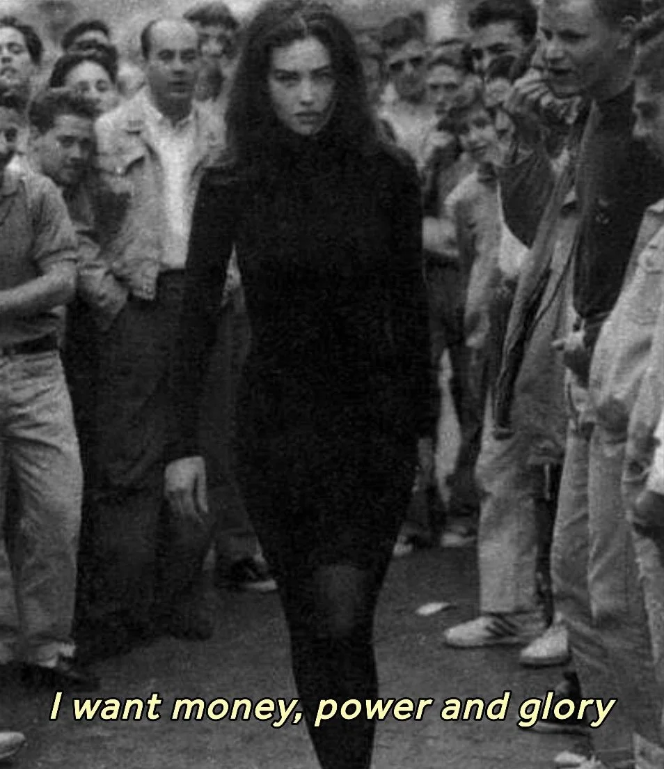 A woman wearing dark clothing walking down a street with a crowd of men on either side watching her, with the caption "I want money, power and glory" at the bottom.