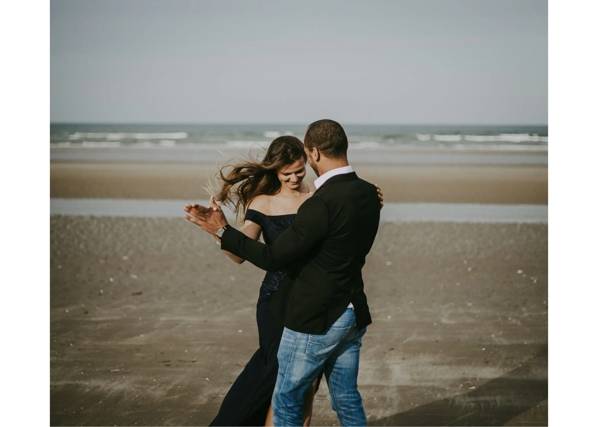 A couple dancing on a beach with waves in the background. The woman has long hair and is wearing a dress, while the man is dressed in a blazer and jeans.