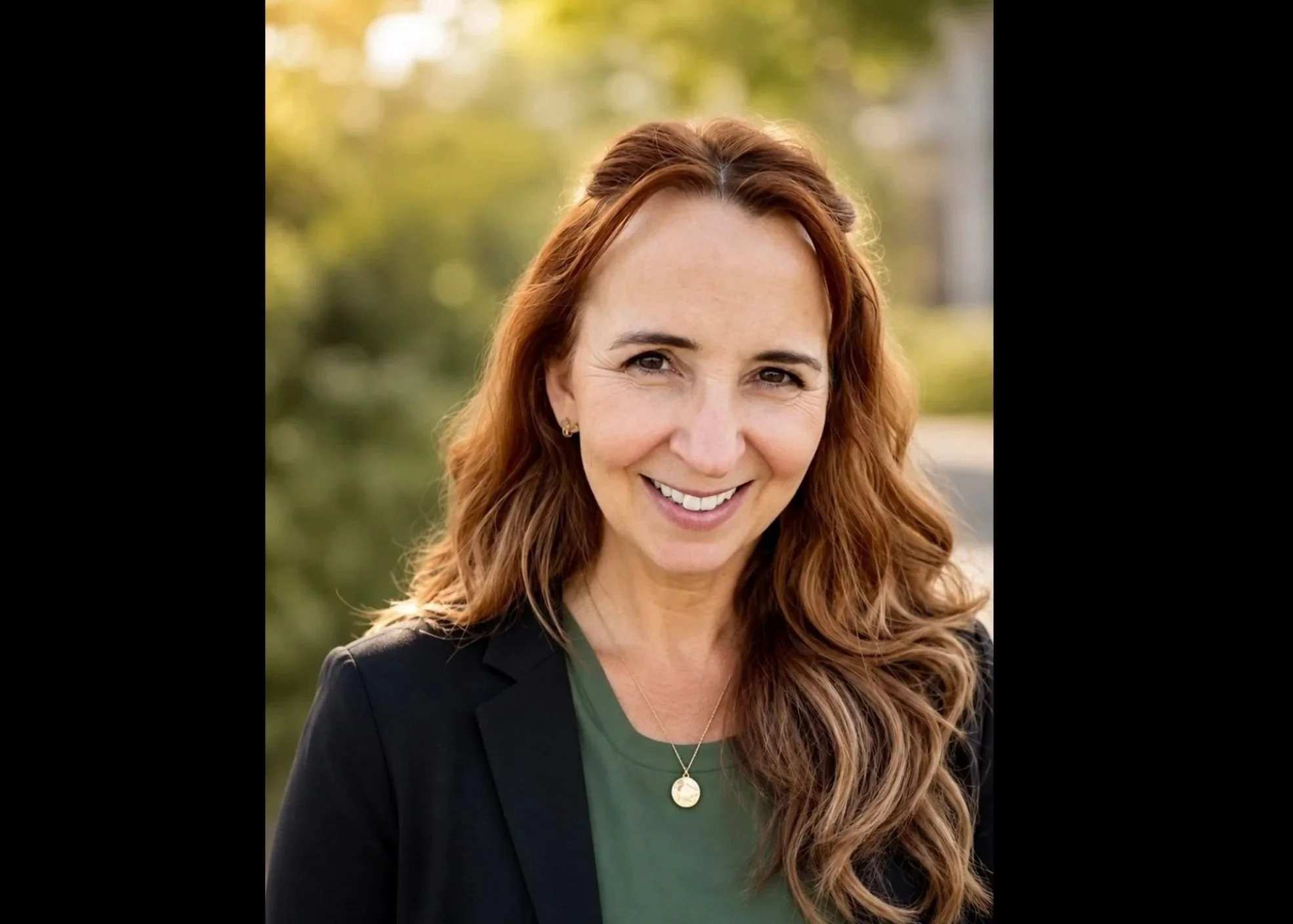 A woman with long red hair, wearing a black blazer and green top, smiling outdoors with a blurred background of trees and sunlight.