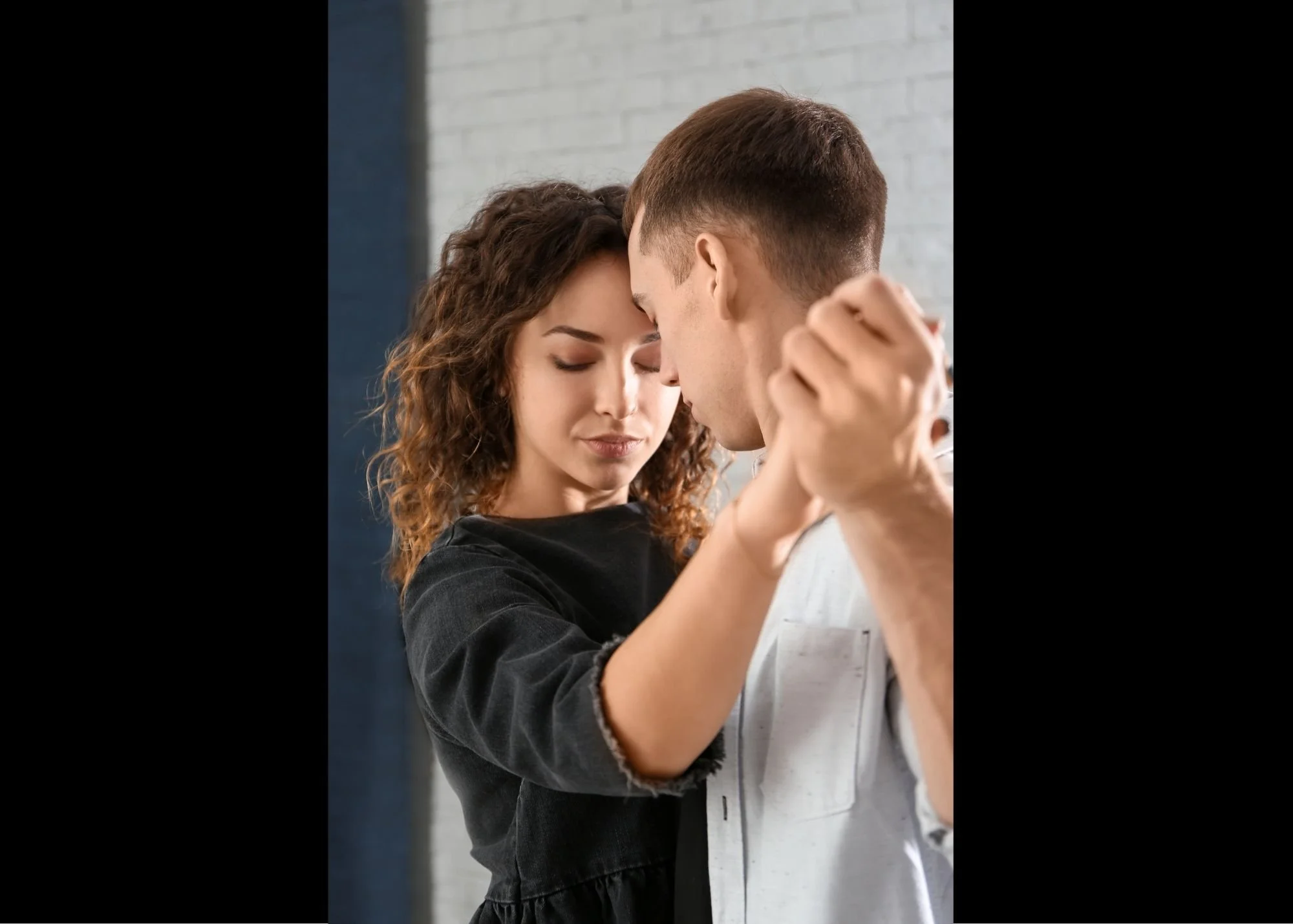 A young man and woman are engaged in a close, intimate dance, with foreheads touching and eyes closed, in a relaxed indoor setting.