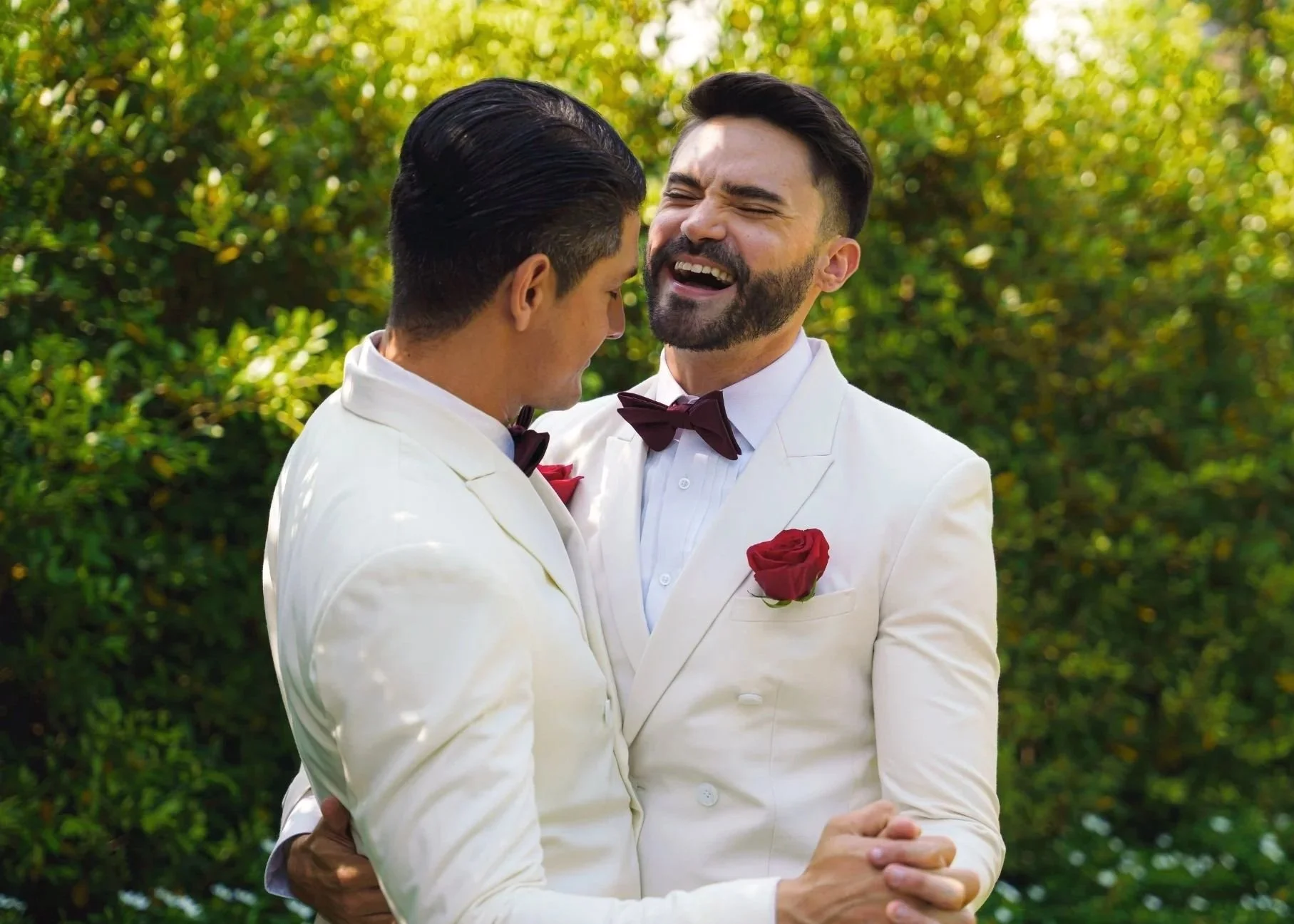 Two men in white tuxedos with red roses on their lapels, embracing and smiling joyfully outdoors with green foliage in the background.