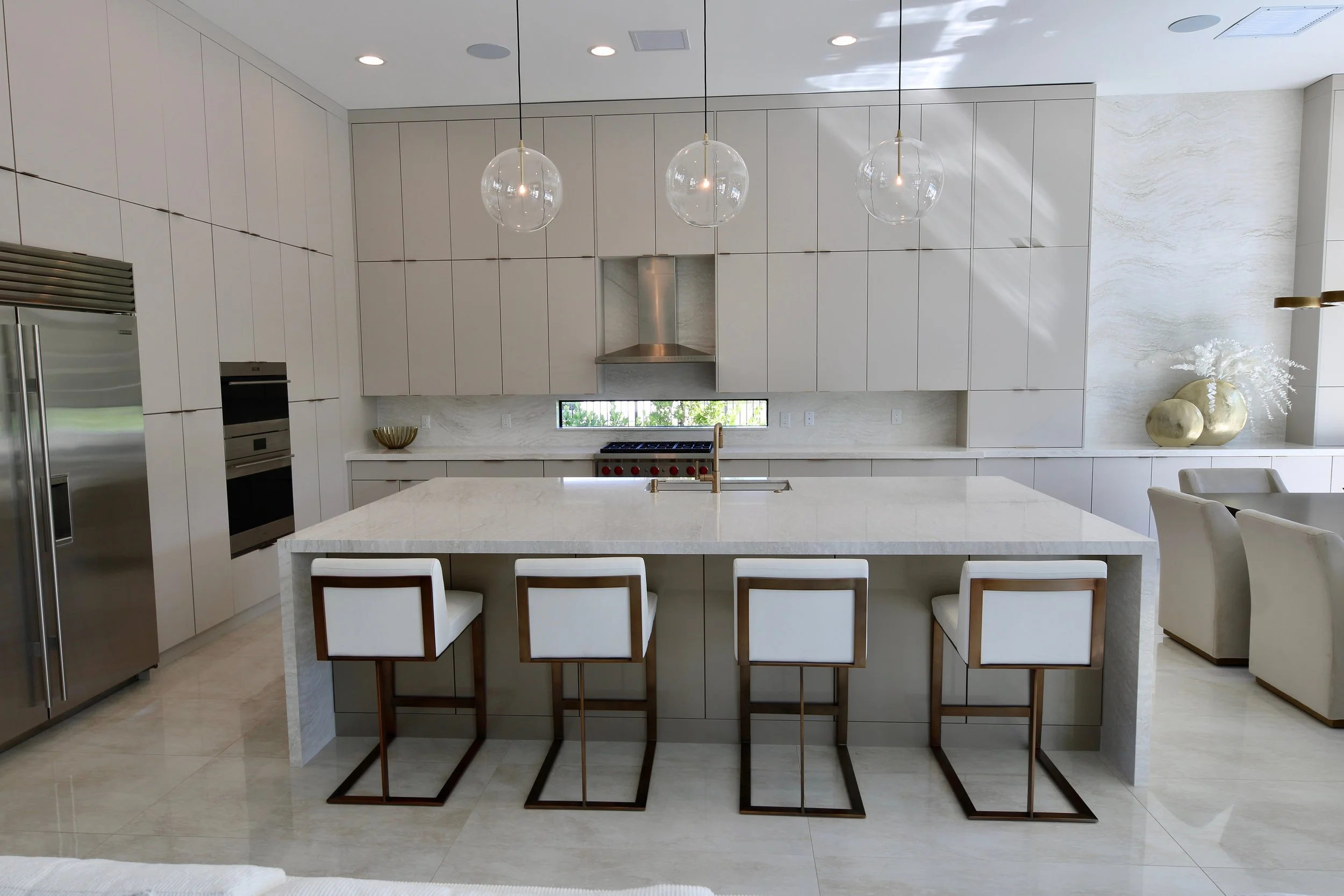 Modern kitchen with white cabinetry, a large island with four chairs, pendant lights hanging above, and a small window behind the stove area. Decor includes a large vase with white flowers and decorative golden vases.