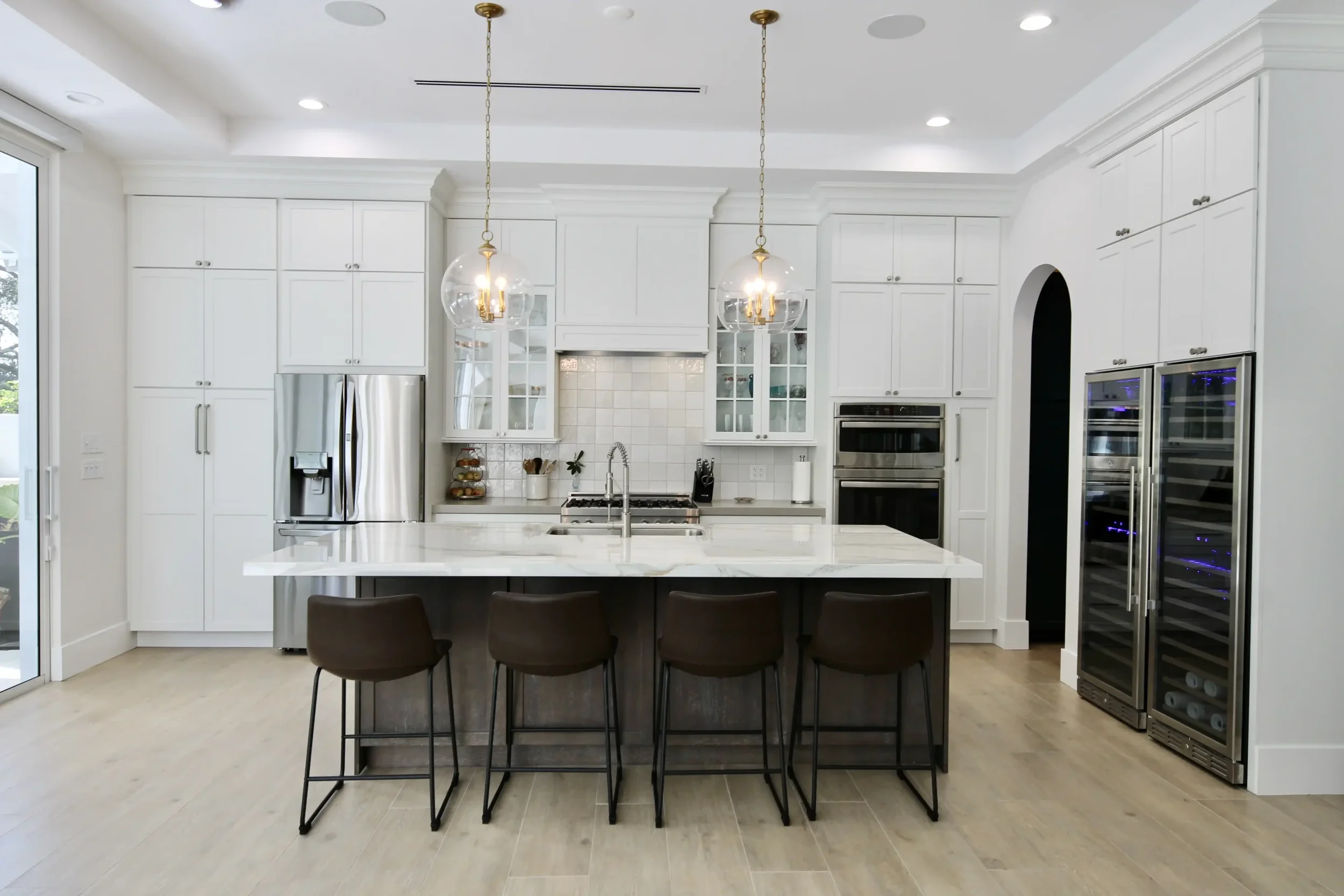 Modern white kitchen with island and black stools, pendant lights, stainless steel appliances, and wine coolers.