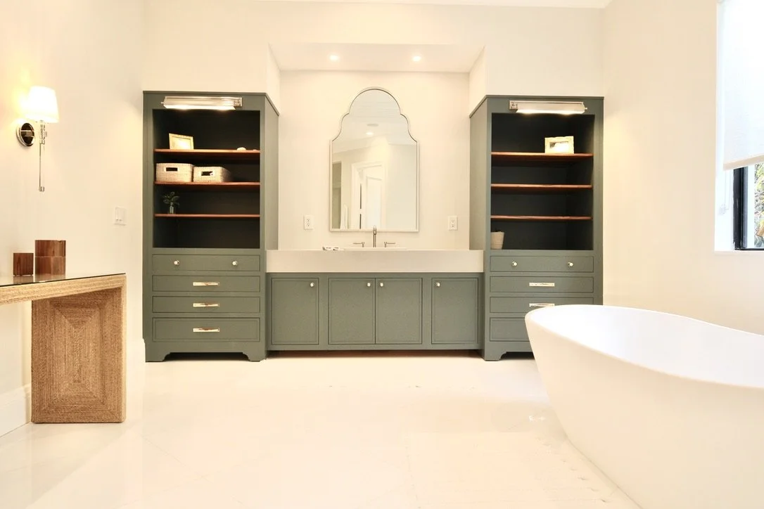 Bathroom with a freestanding bathtub on the right, a light blue vanity with a mirror above in the center, and two matching light blue shelving units on either side, with a wooden side table on the left and a window on the right.