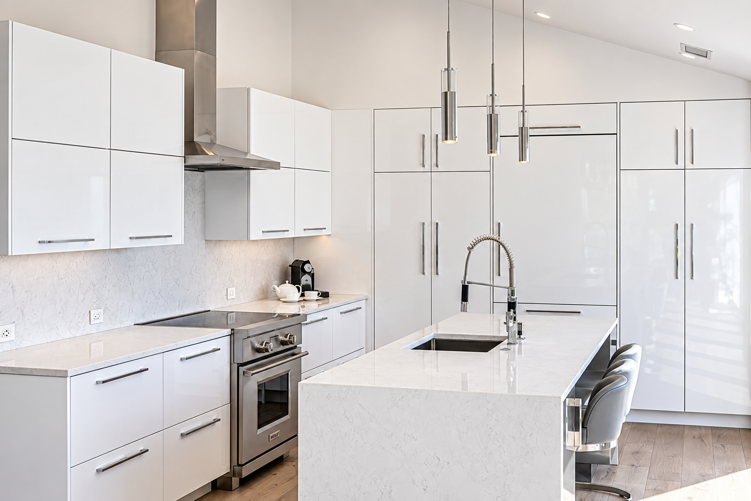 Modern white kitchen with stainless steel appliances, including a stove and oven, a large kitchen island with a built-in sink and chrome faucet, white cabinets, pendant lights hanging above the island, hardwood flooring, and a coffee machine on the c