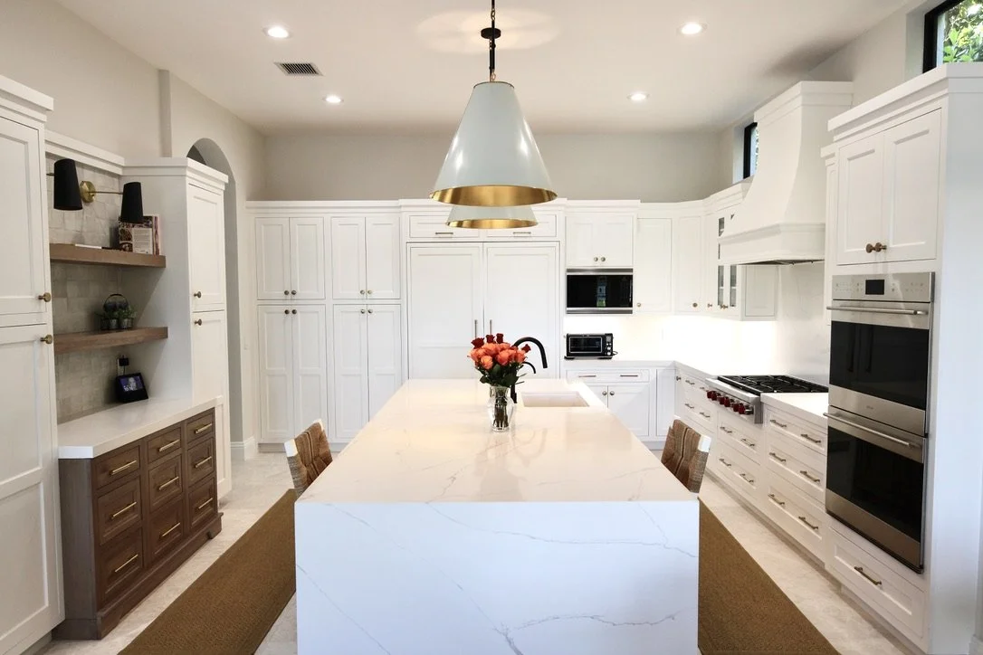 Modern kitchen with white cabinets, marble island, black faucet, bouquet of pink flowers, built-in oven and microwave, stovetop, and wooden chairs.
