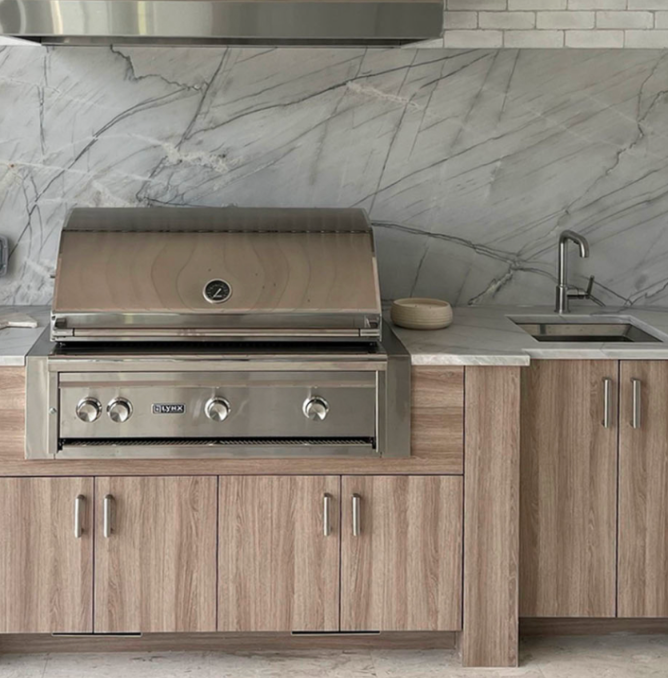 Modern kitchen with stainless steel grill, wood cabinets, marble backsplash, small beige dish, and sink with chrome faucet.