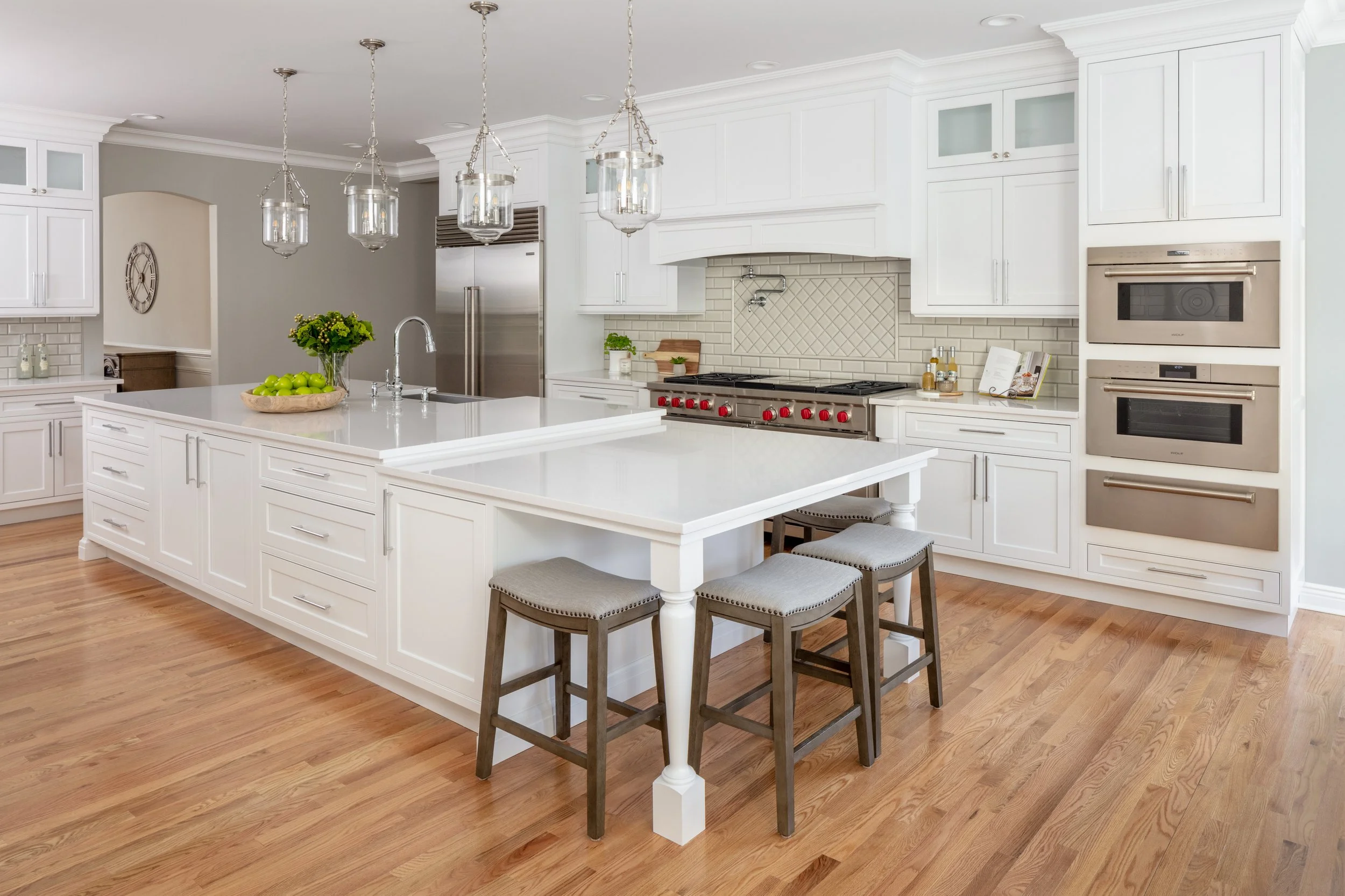 A bright, spacious kitchen with white cabinets, a large island with a white countertop, three hanging pendant lights, stainless steel appliances, including a double oven and a refrigerator, and hardwood flooring. Decor items include a bowl of green a