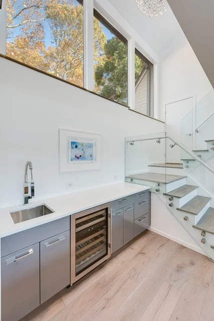 Modern kitchen area with gray cabinets, a built-in wine cooler, a white countertop with a sink, large windows showing trees outside, and a staircase with glass railing.