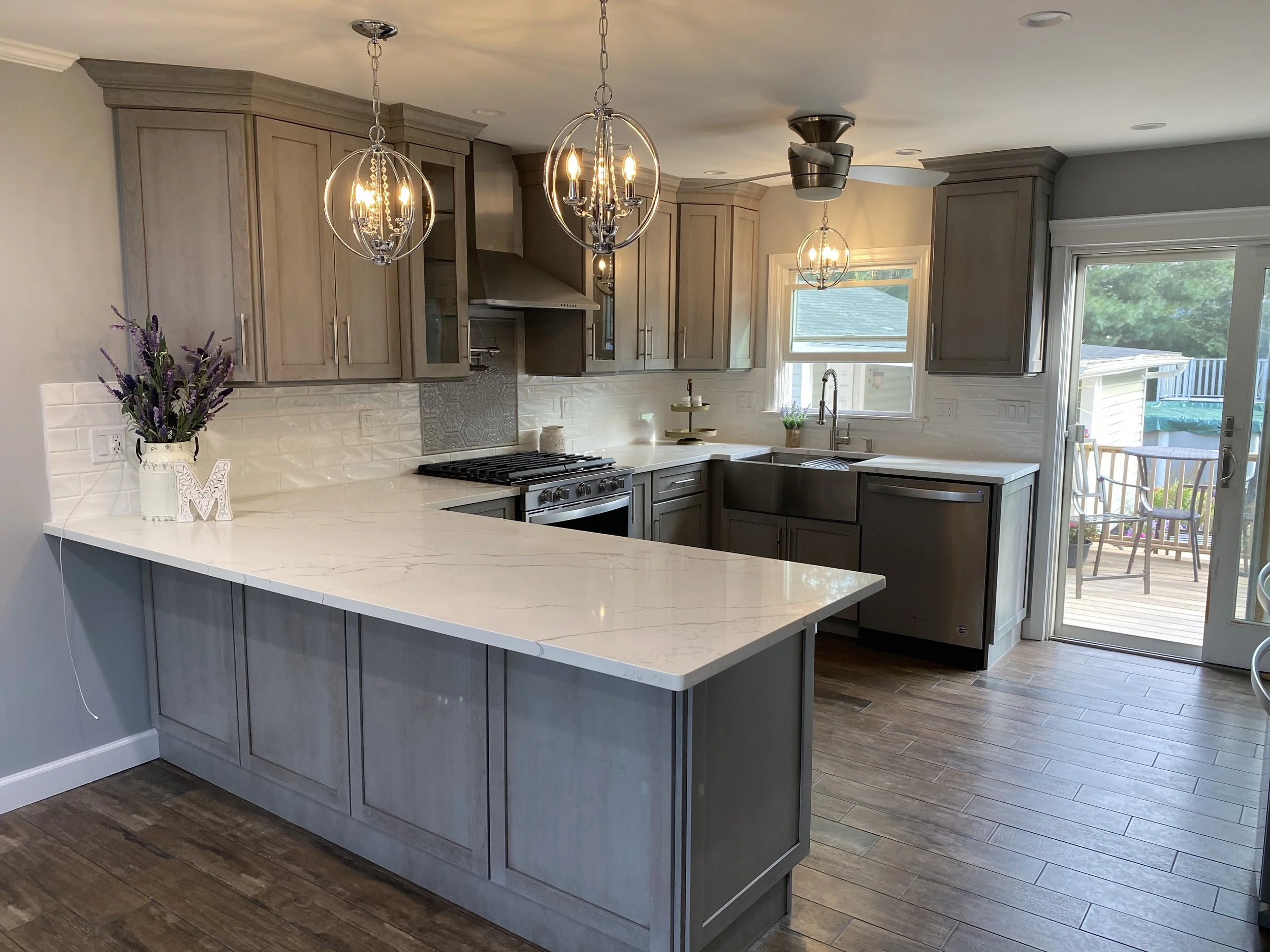 Modern kitchen with gray cabinets, white marble countertops, pendant lights, a stove, and a window overlooking a backyard with a deck and outdoor furniture.