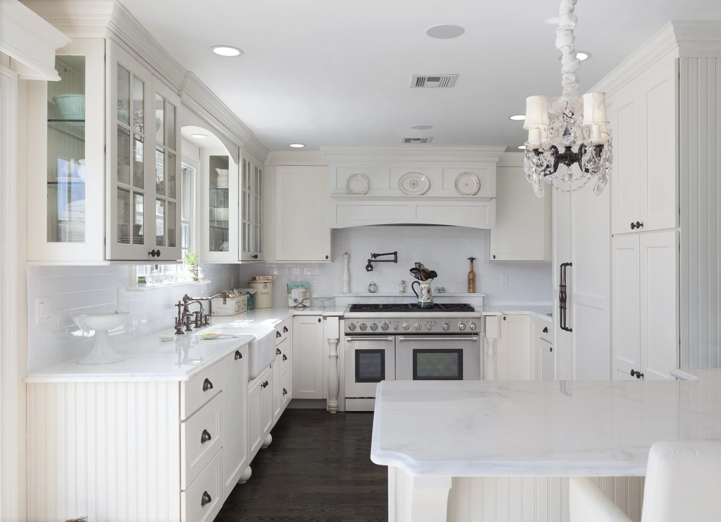 A bright, white kitchen with glass-front cabinets, a white marble island, and a chandelier hanging from the ceiling.