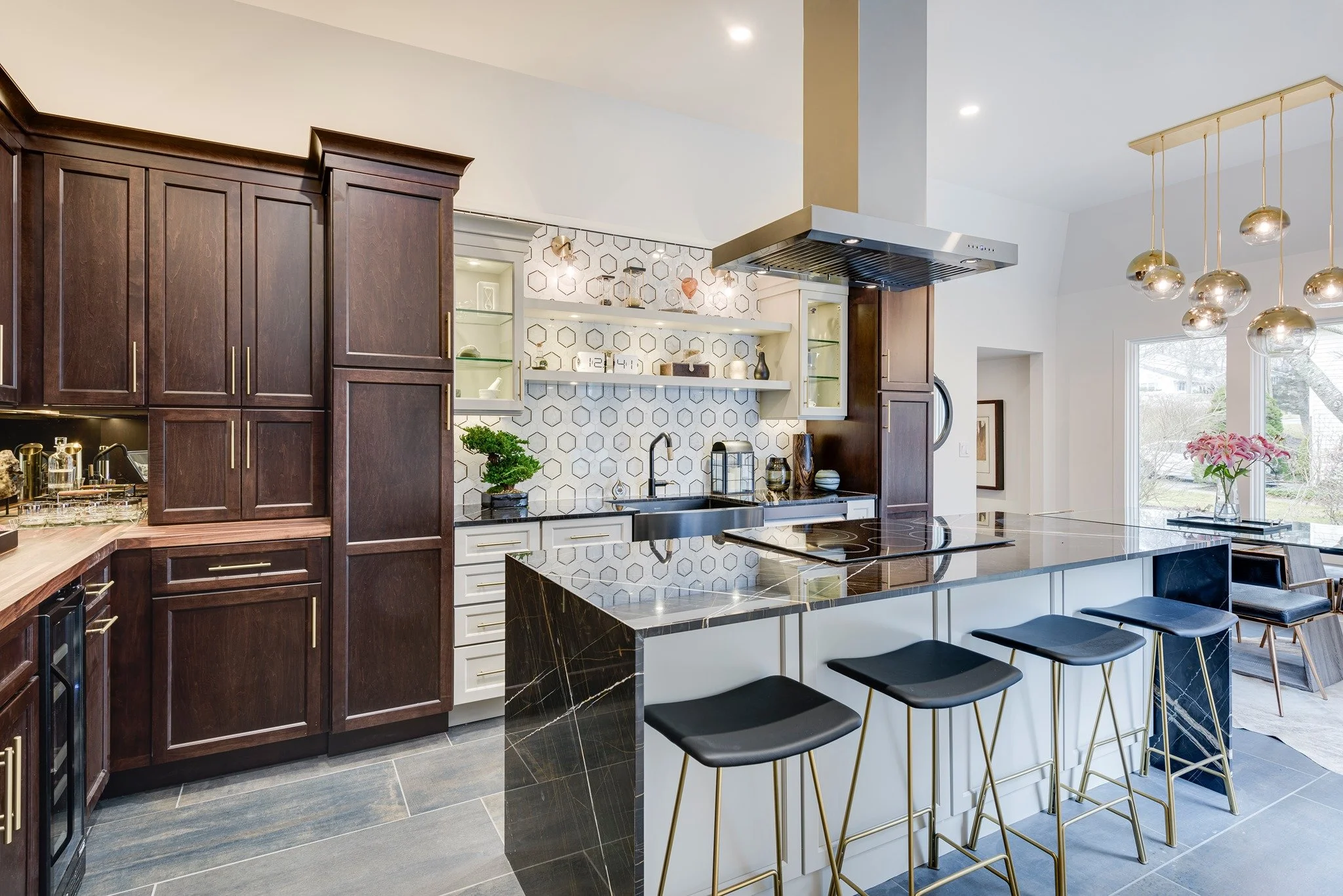 Modern kitchen with dark wood cabinets, black marble island, white shelves with decorative items, gold pendant lights, and a large window featuring a pink flower arrangement.