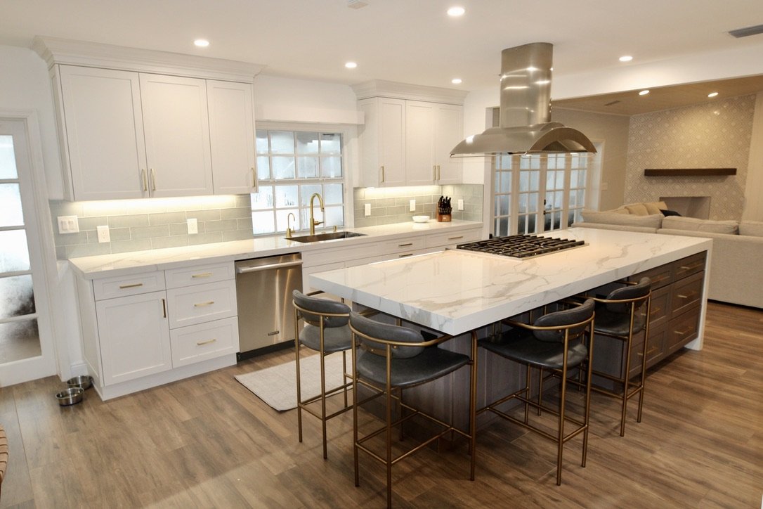 Modern white kitchen with an island, marble countertop, stainless steel appliances, and bar stools, adjacent to a living area.