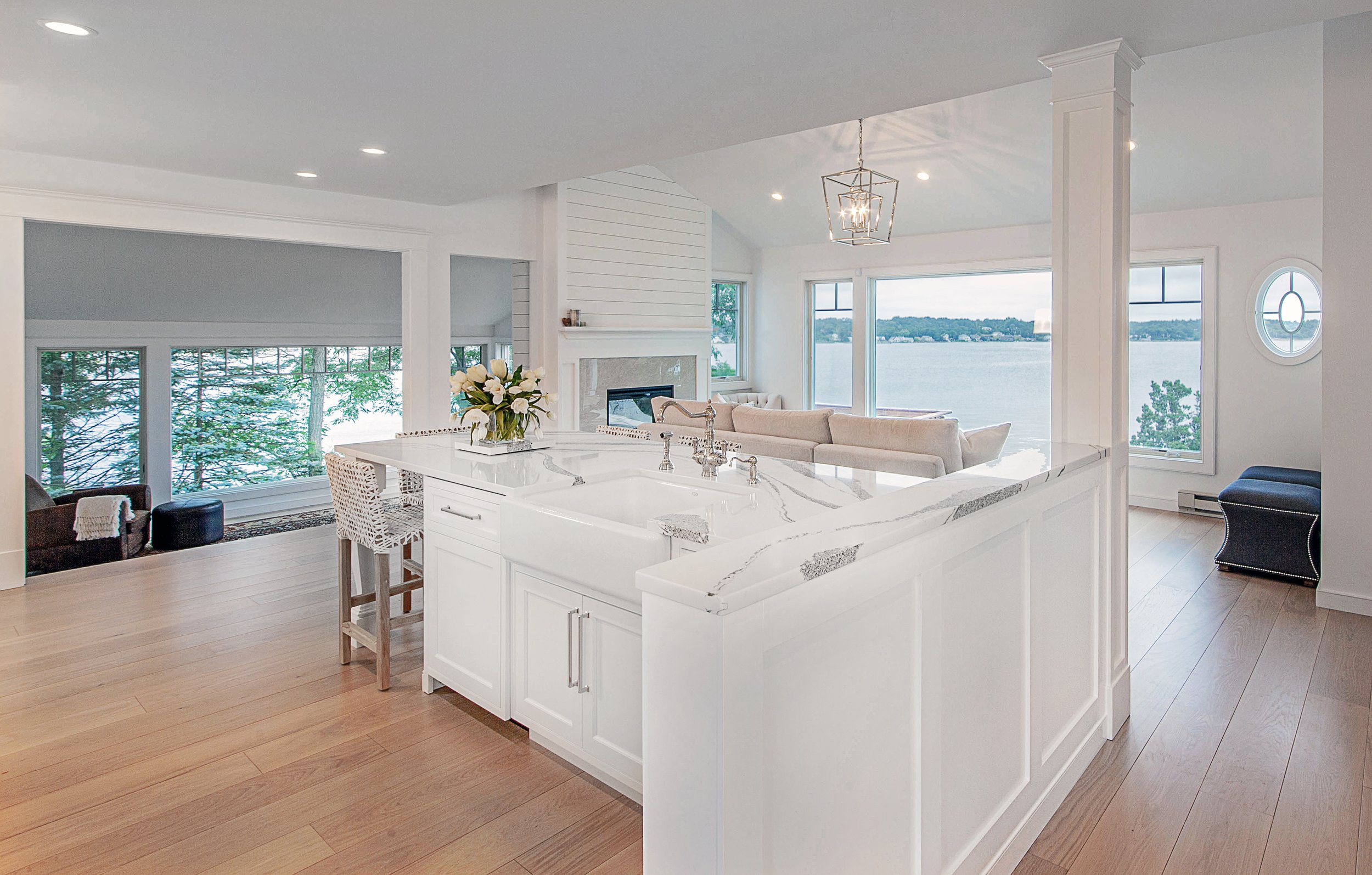 Open-concept living room and kitchen with large windows overlooking water, white cabinets, marble kitchen island, hardwood floors, modern chandelier, and cozy seating areas.