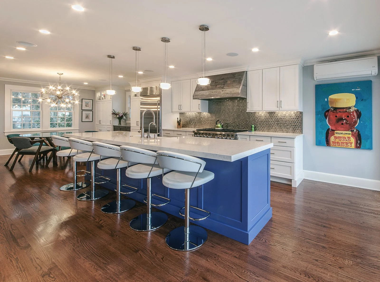 Modern kitchen with white cabinets, a large island with a blue base, white bar stools, a stainless steel stove, and a colorful artwork on the wall.