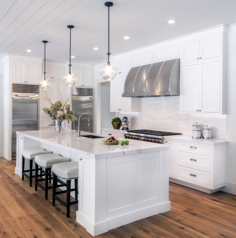 Modern white kitchen with a large island, pendant lights, stainless steel appliances, and wooden floors.