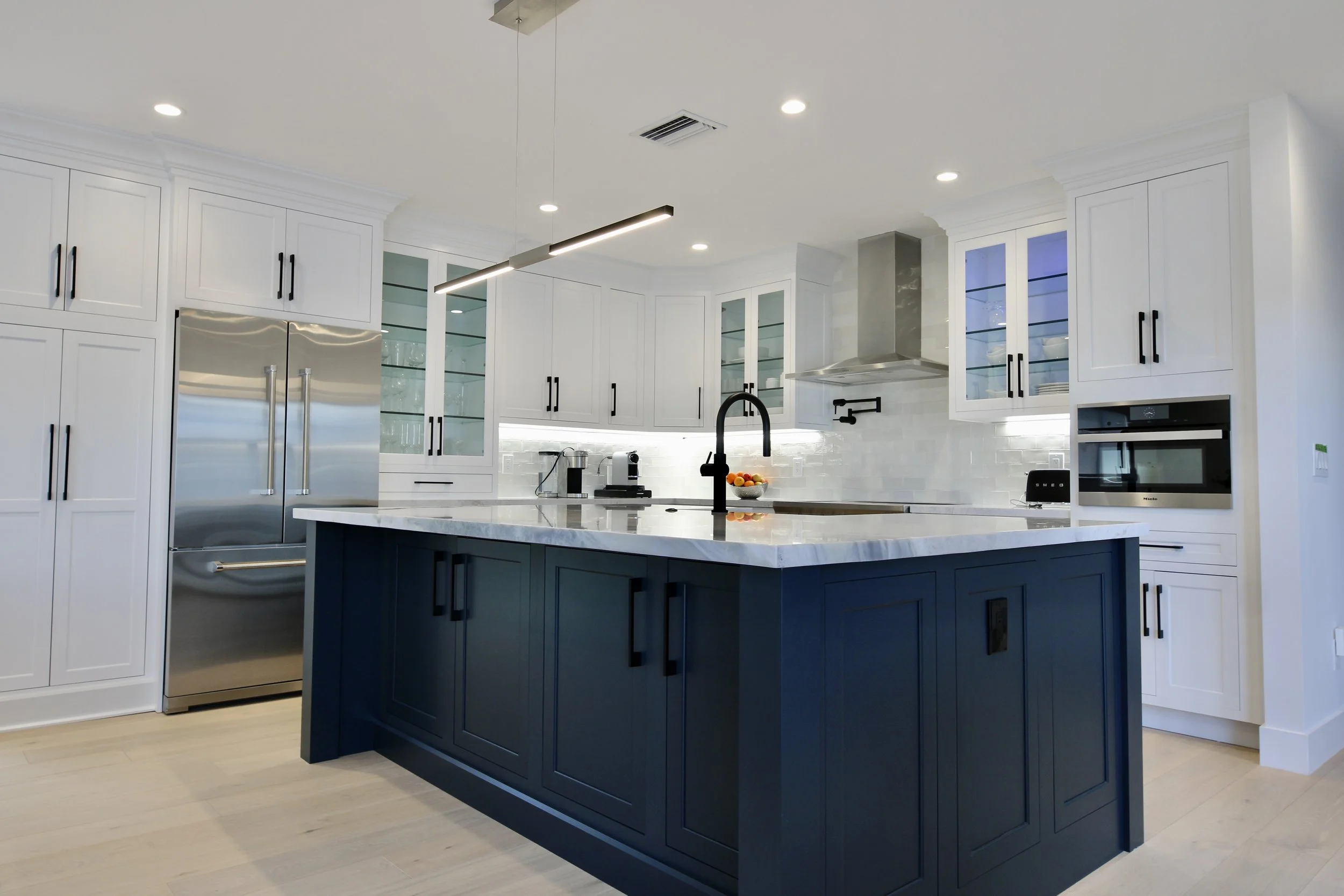Modern kitchen with white cabinets, a navy island, and stainless steel appliances, including a refrigerator and an oven, under bright recessed lighting.