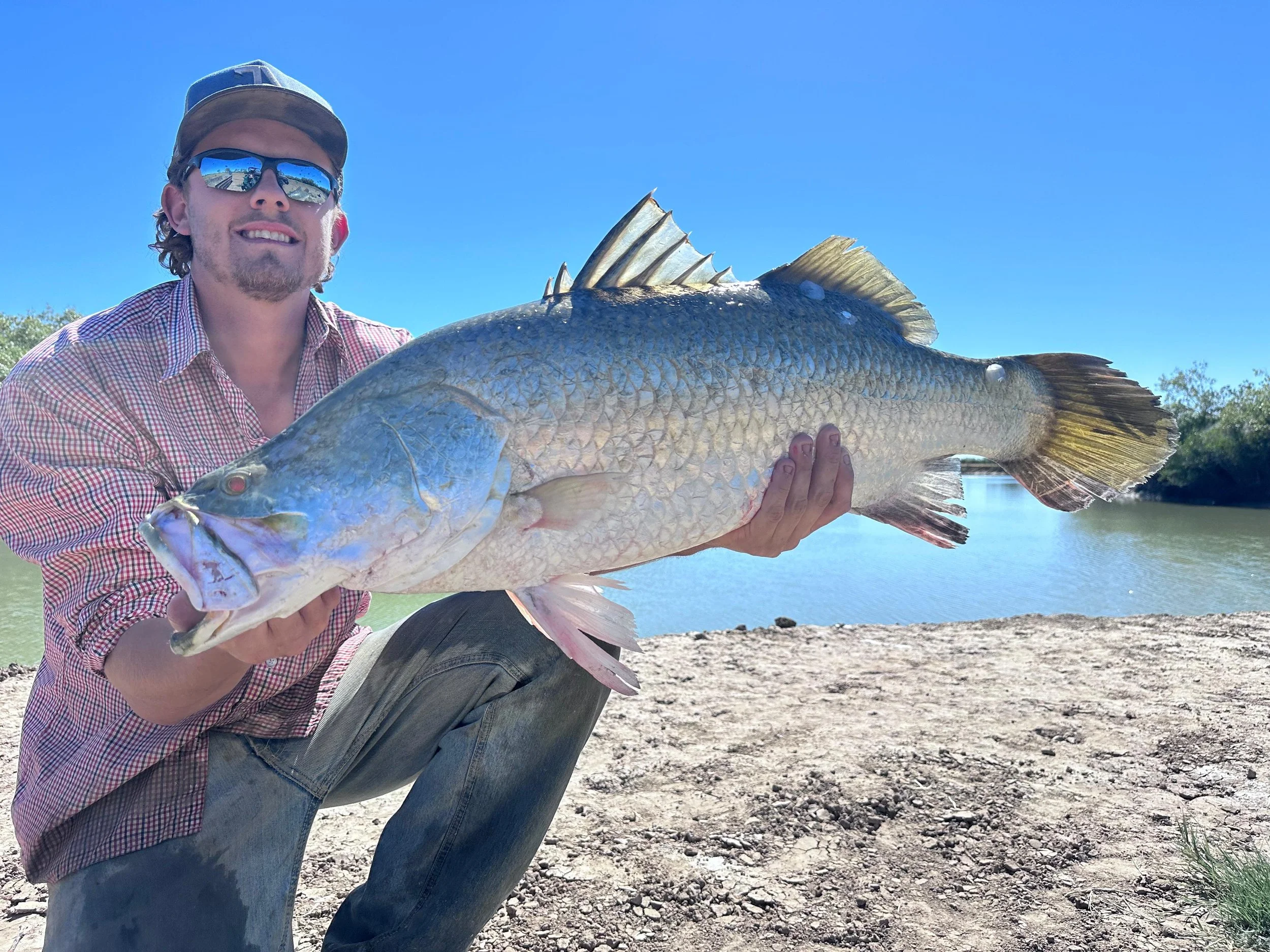 A man wearing sunglasses, a baseball cap, and a checkered shirt holding a large fish beside a body of water on a sunny day.