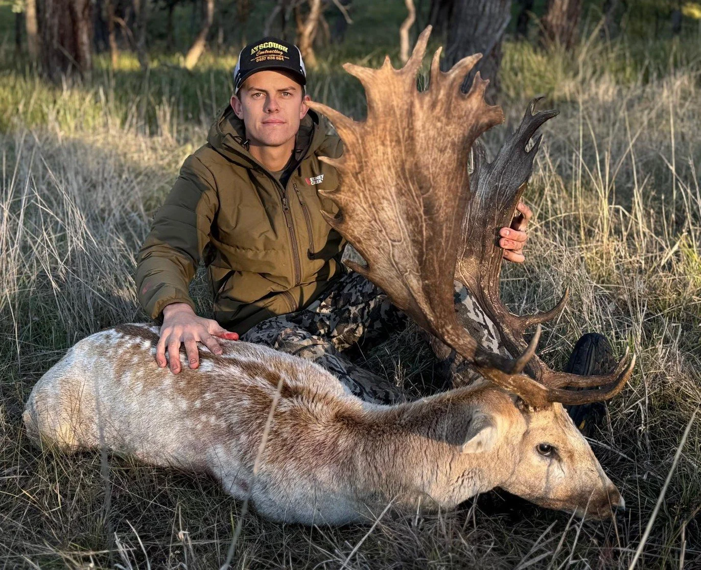 A man hunting outdoors sitting in grass with a large deer with antlers, holding its head and resting his hand on its body.