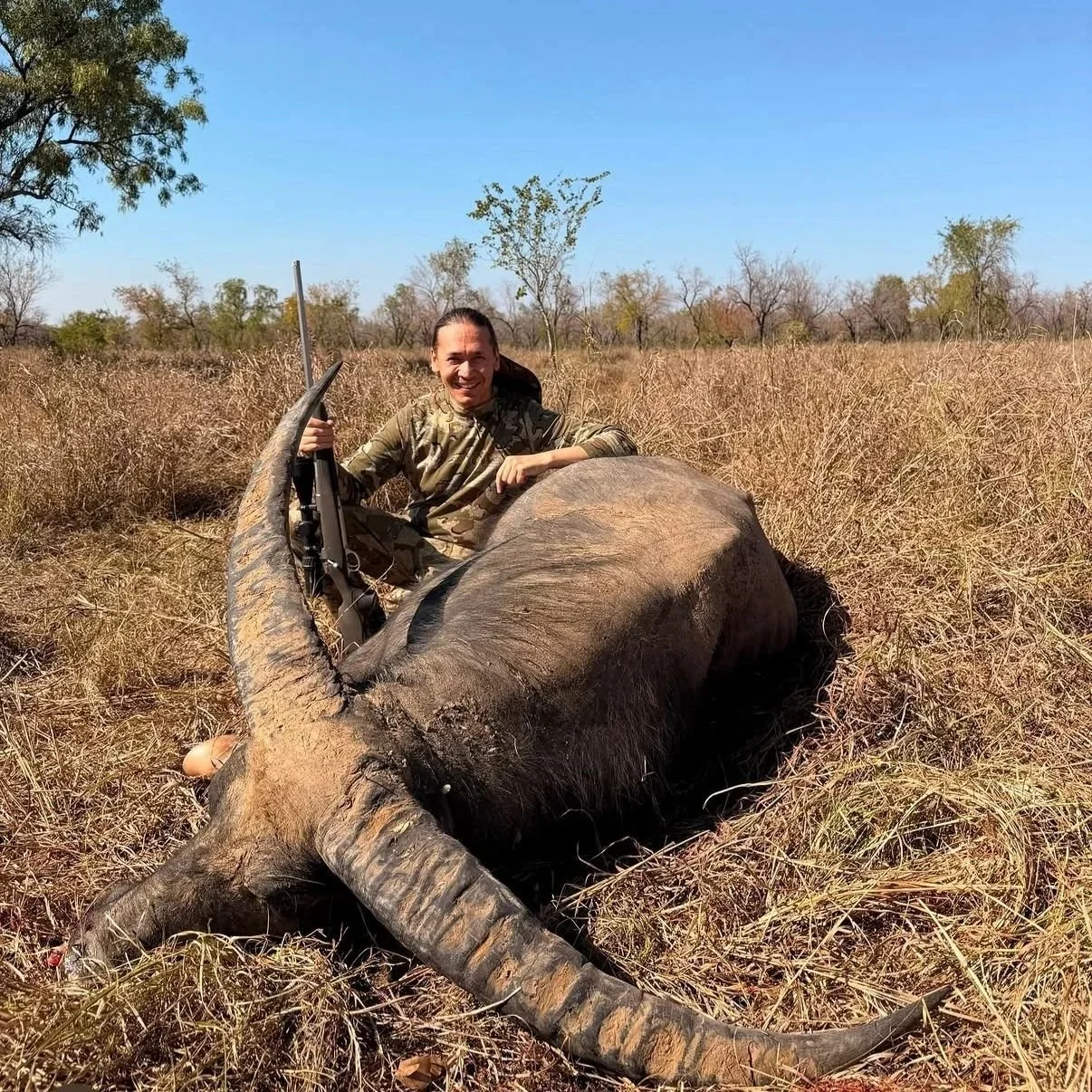 A person in camouflage clothing sitting on a dead elephant lying in dry grass in a savanna landscape, holding a rifle, with a smile on their face.