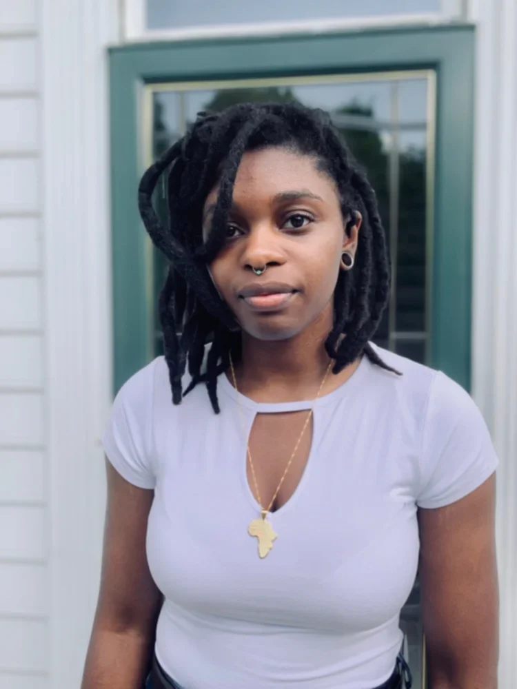 A young woman with dreadlocks standing outside in front of a door, wearing a light-colored shirt and a gold necklace with an Africa-shaped pendant.