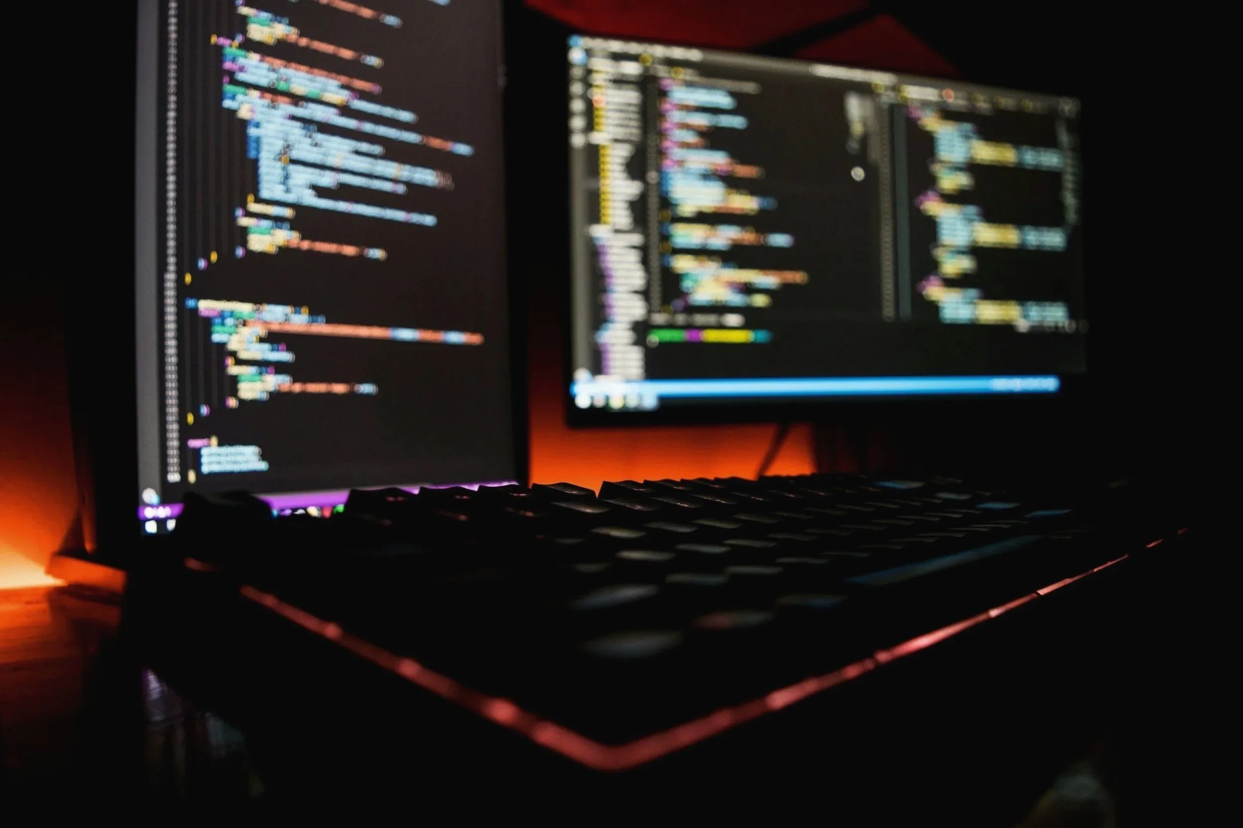 Two computer monitors displaying lines of colorful code in a dark room with orange backlight lighting.