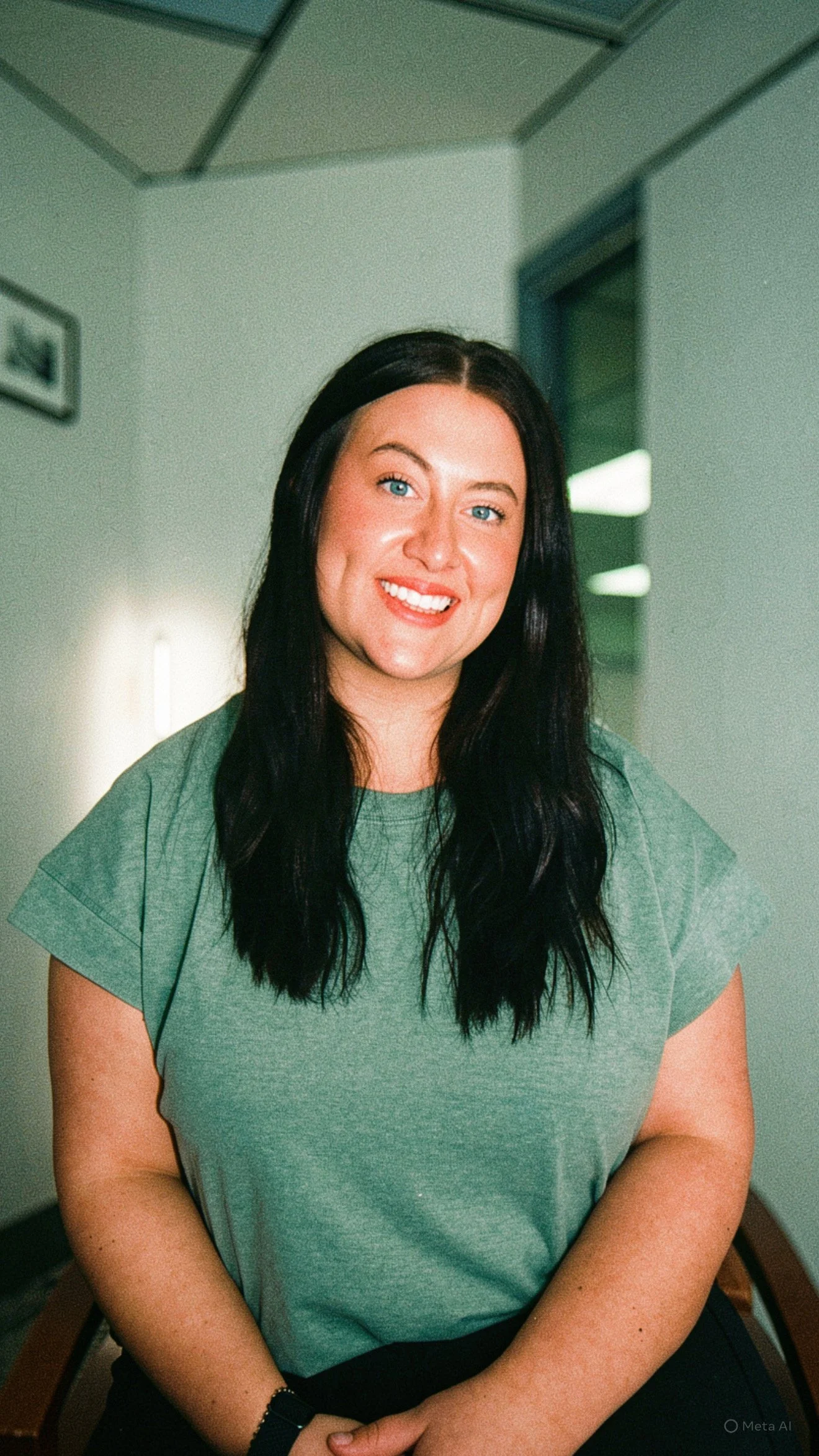 Young woman with black hair and blue eyes sitting indoors in front of a lighted background, wearing a green t-shirt and smiling.