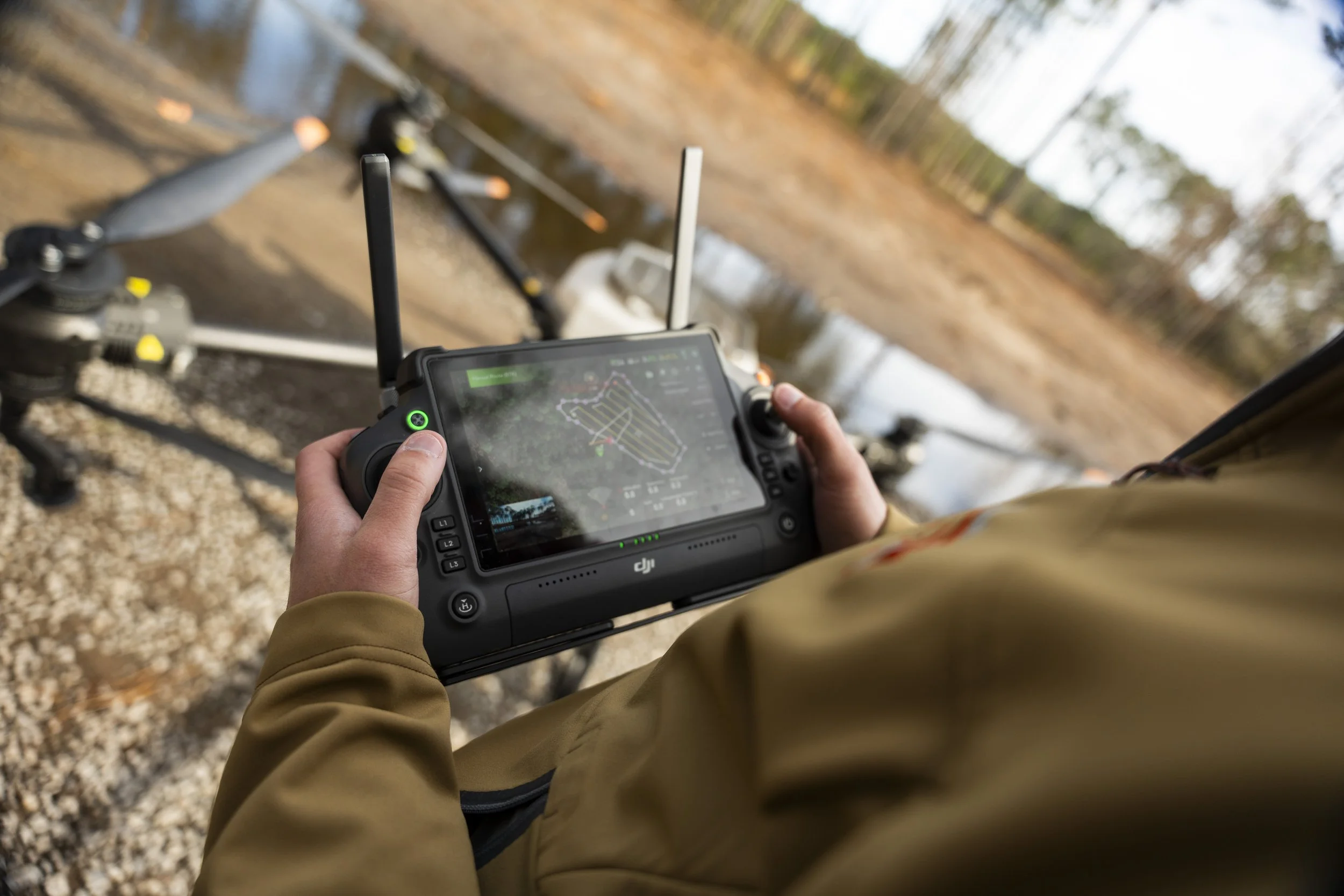 Person holding a drone remote controller with a drone flying overhead in an outdoor wooded area.
