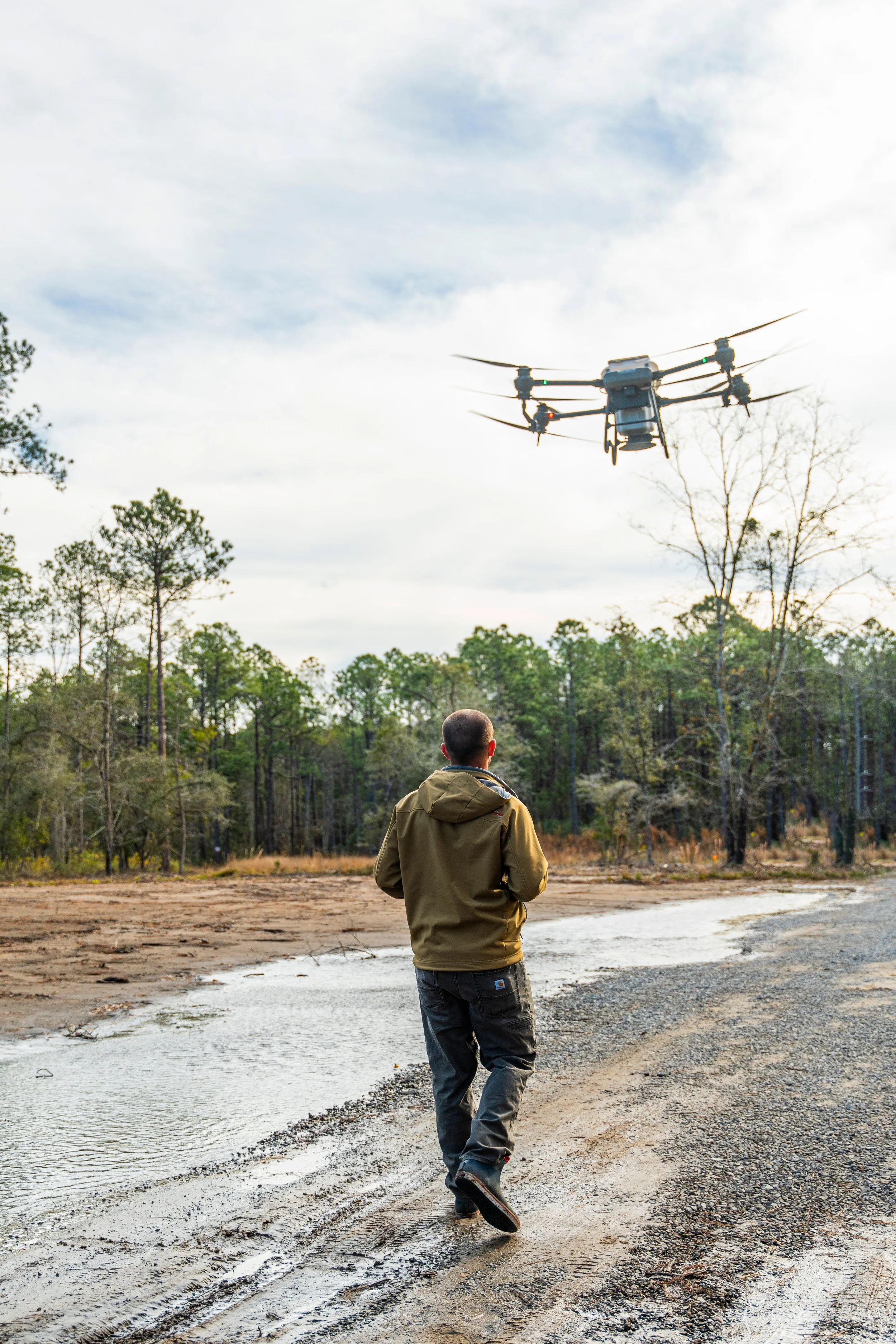 A man in a brown jacket and jeans walking on a muddy trail near a small body of water, with a drone flying overhead in a forested area with tall trees and a cloudy sky.