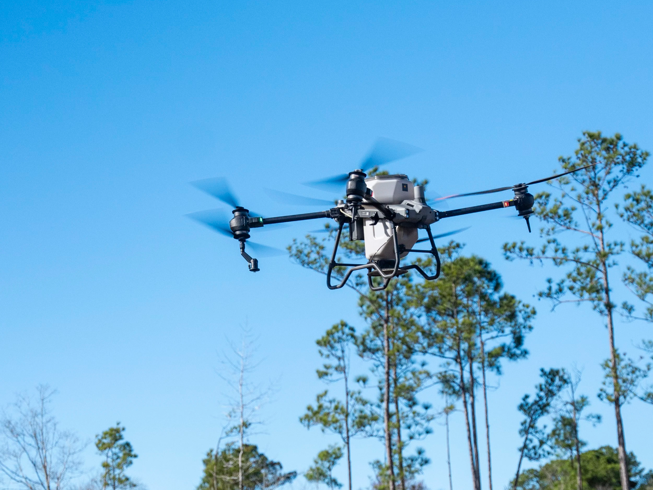 A drone flying outdoors in a clear blue sky with green trees in the background.