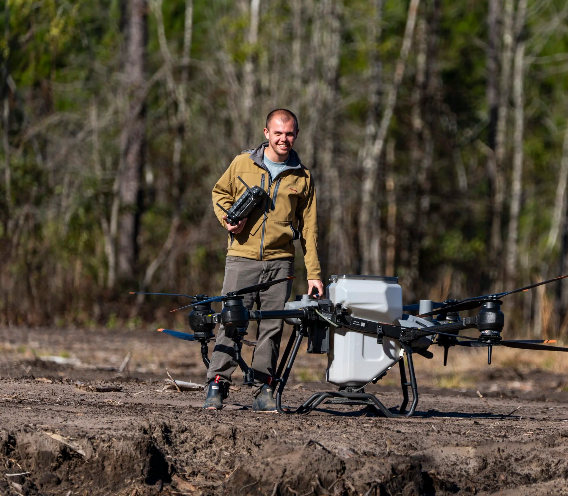A man outdoors in a wooded area, smiling and holding a remote control, standing next to a large drone with cameras and equipment.