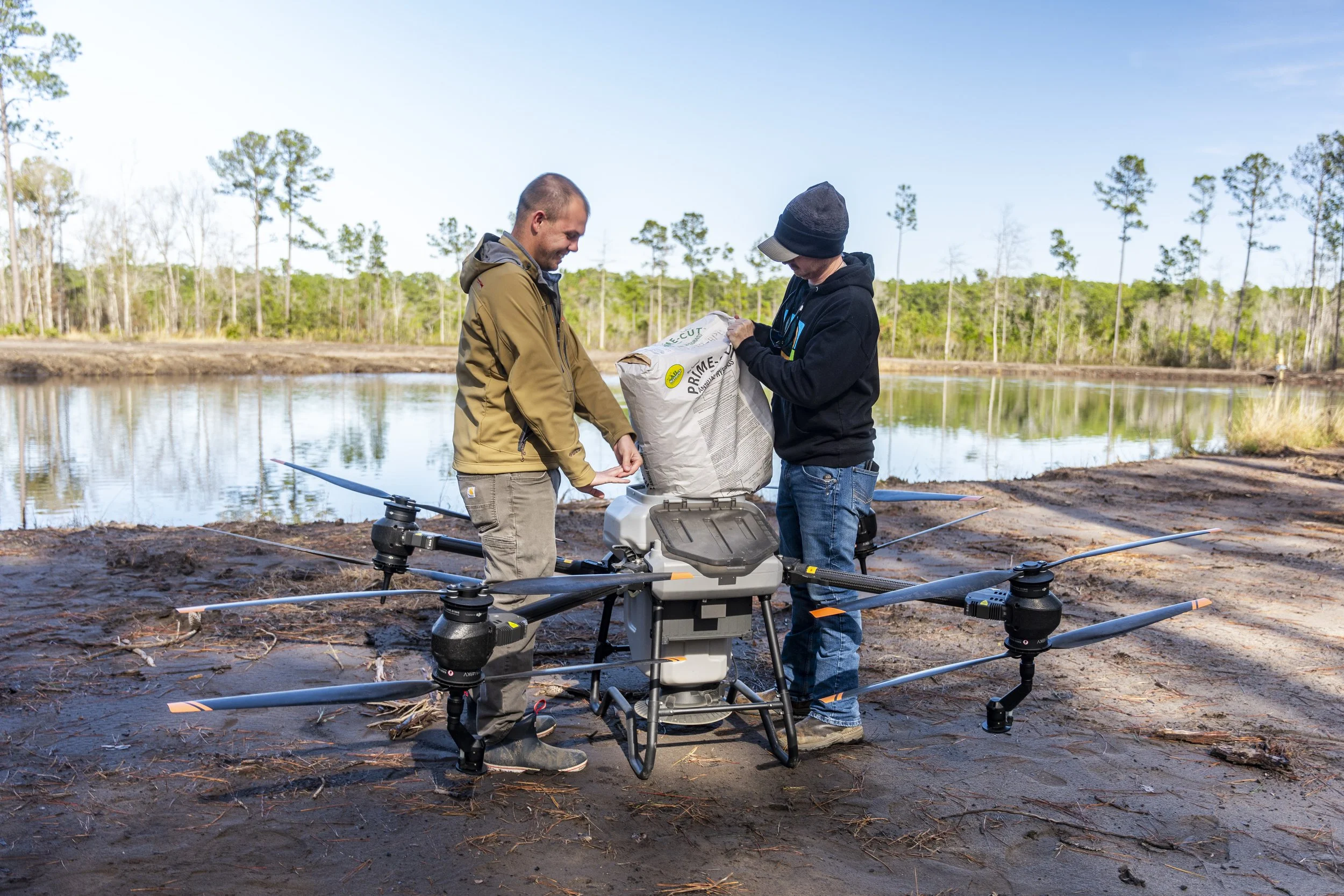 Two men preparing a drone with a package near a lake in a forested area, with trees and water in the background.