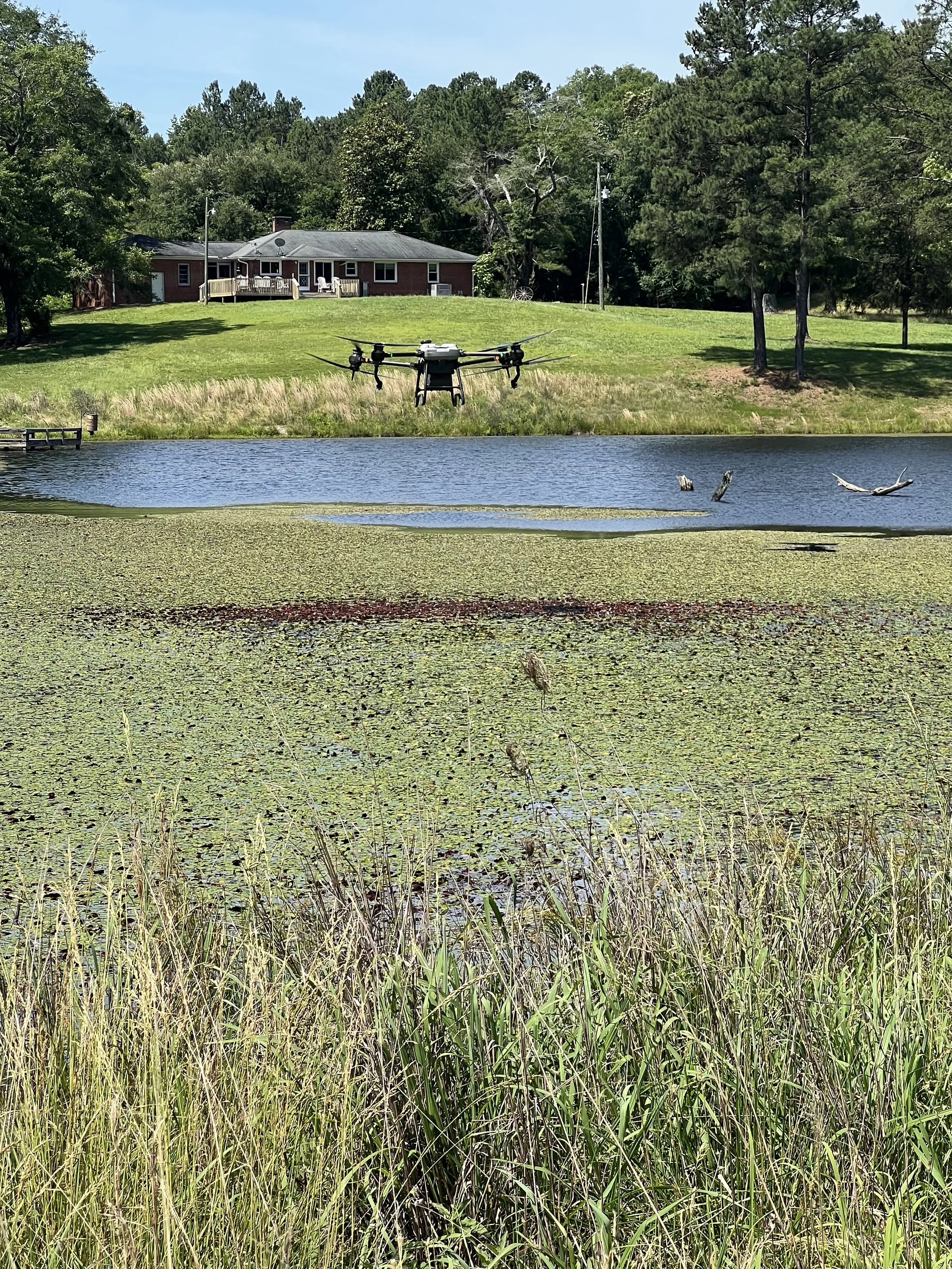 A drone flying over a pond near a grassy yard with a house and trees in the background.