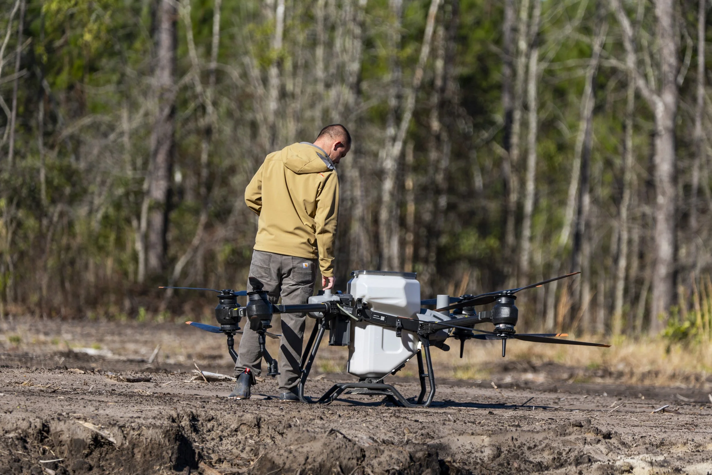 A man in a tan jacket and gray pants is working with a large drone in a wooded outdoor area.