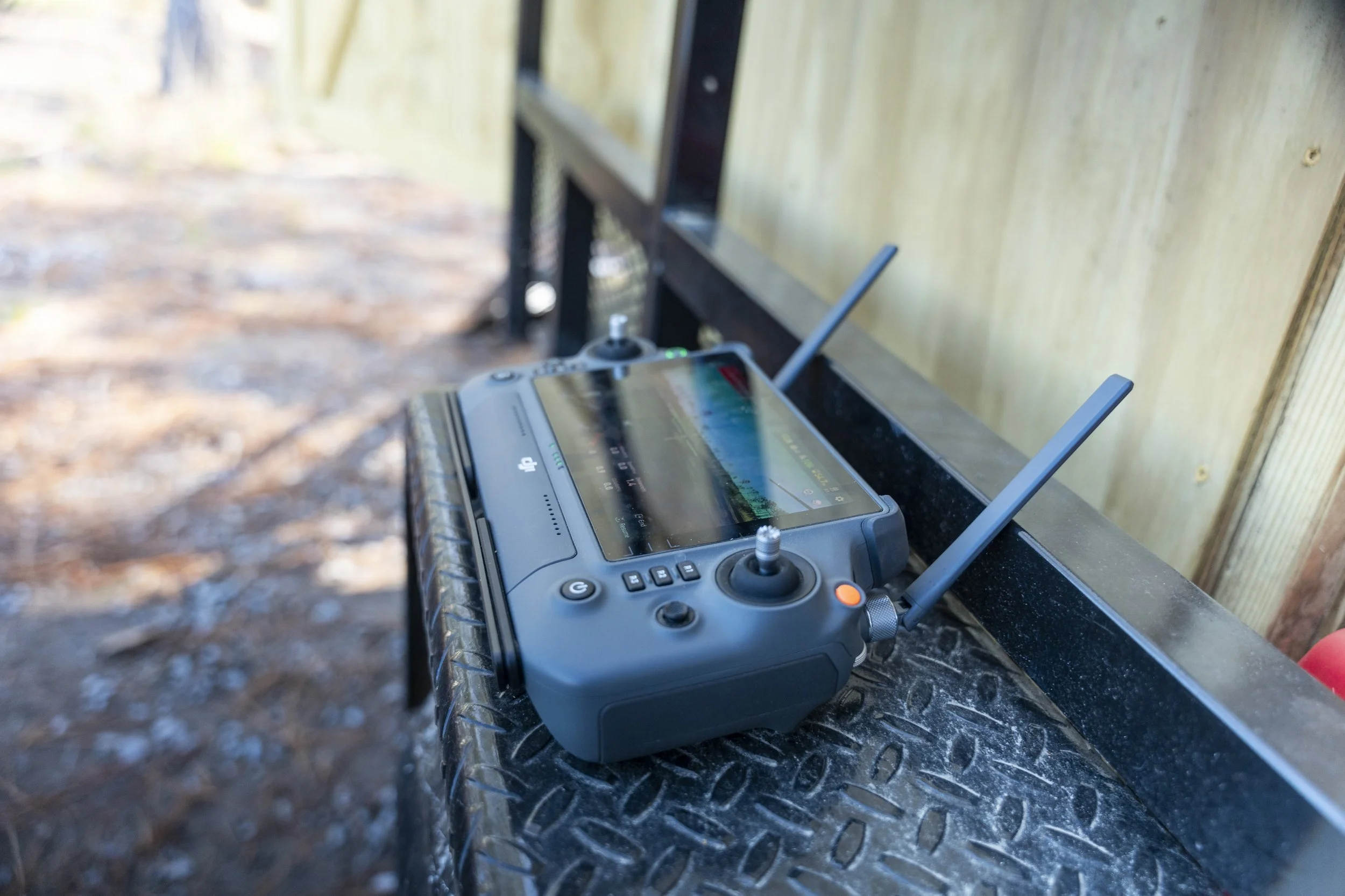 A remote control device with antennas and a display screen resting on a black textured surface outdoors, with a wooden structure in the background.