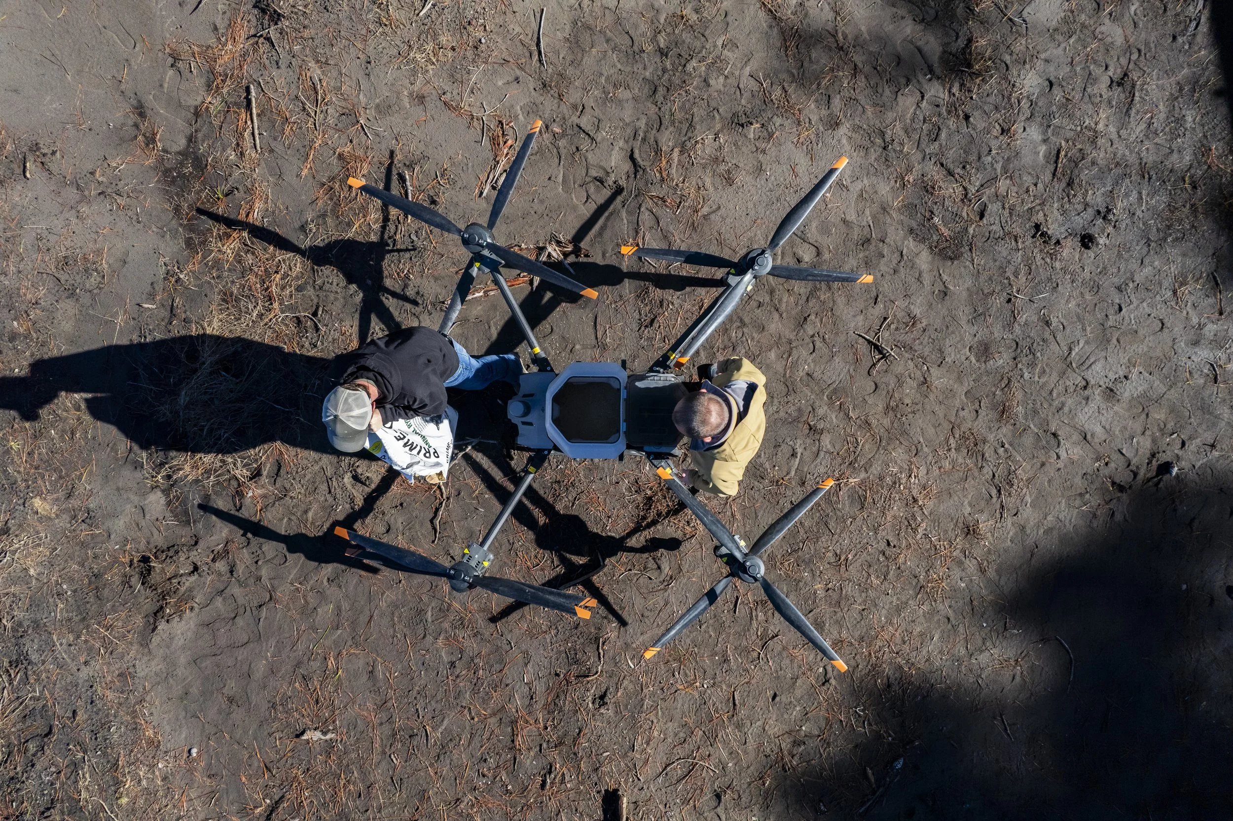 Bird's eye view of two men working with a piece of scientific or surveying equipment on a patch of barren ground with dry leaves and twigs. One man is wearing a black jacket and a beige cap, while the other is in a yellow jacket with a hood. Their shadows are cast on the ground.